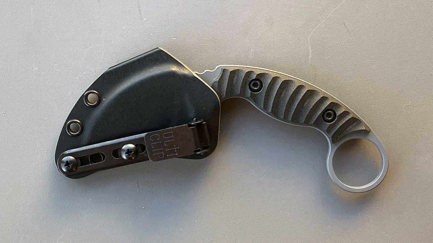 Image of the Toor Knives Karsumba R beside its custom-molded Kydex sheath. The sheath features mounting holes and retention adjustments for different carry styles, from belt clips to static line setups. The design reflects Toor’s focus on practical, real-world usability rather than flashy presentation. The knife’s curved blade and retention ring pair cleanly with the sheath’s form-fitting profile. This photo supports the review’s discussion of how the Karsumba R balances secure retention with smooth deployment. It emphasizes the knife’s role as a purpose-built defensive tool with a professional-grade sheath.