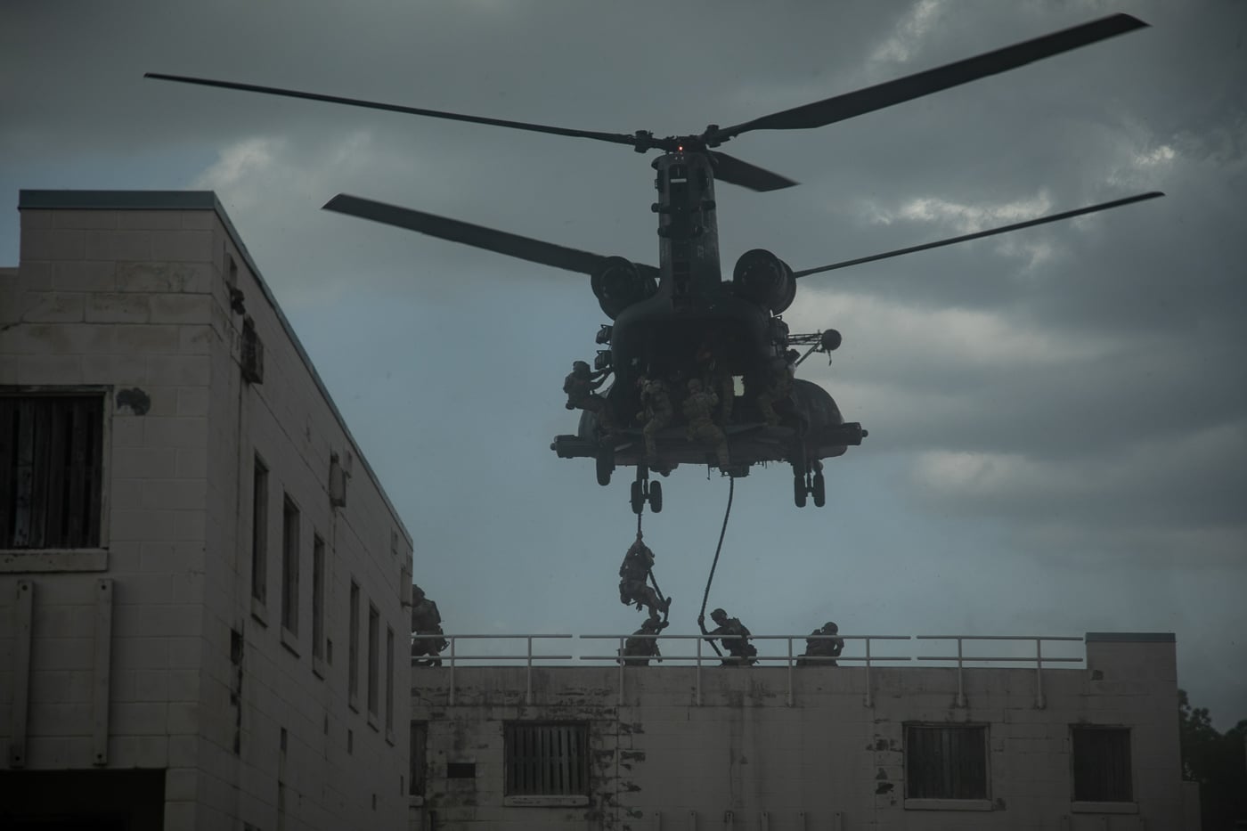 The image shows U.S. Army Rangers assigned to the 75th Ranger Regiment fast-roping from the rear ramp of a 160th Special Operations Aviation Regiment (Airborne) MH-47 Chinook helicopter during an airborne assault demonstration at CAPEX2025, held at Fort Bragg. The helicopter is hovering just above the ground with its rear ramp lowered and thick ropes extending down, as Rangers in full tactical gear and helmets rapidly descend onto a grassy landing zone. Spectators and additional military personnel may be visible in the background, watching the demonstration. The Chinook’s distinctive twin rotors and dark Night Stalkers livery are prominent. This scene highlights the advanced insertion techniques practiced by elite special operations forces, demonstrating their ability to deploy quickly and efficiently from rotary-wing aircraft. The demonstration showcases the partnership between the 160th SOAR and the 75th Ranger Regiment, as well as the high standards of training maintained by both units.