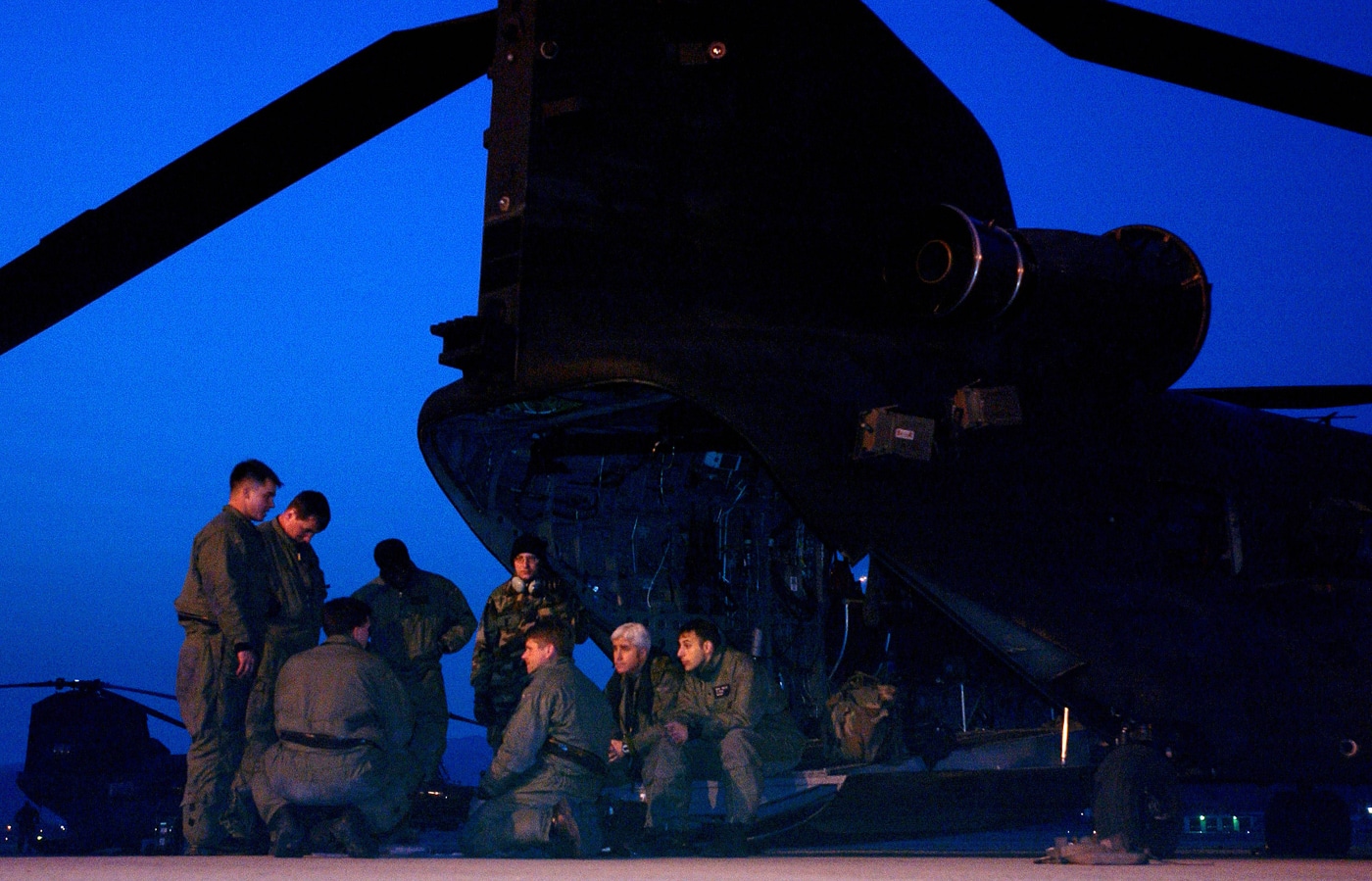 The image shows US Army personnel assigned to the 160th Special Operations Aviation Regiment (Airborne) gathered around an MH-47 Chinook helicopter on the ground in Korea. The crew is receiving a pre-flight briefing from a mission leader, who is pointing to a map or checklist as the group listens attentively. The Chinook’s rear ramp is down and the aircraft is parked on a tarmac or staging area, with additional support equipment and vehicles nearby. This pre-mission briefing is taking place in support of exercises Foal Eagle and Reception, Staging, Onward Movement and Integration (RSOI), both of which are major joint and combined military drills. The scene emphasizes teamwork, communication, and detailed planning before launch. Personnel are in flight suits or operational uniforms, with helmets and other mission gear visible. The image highlights the complexity and coordination required for large-scale multinational operations involving special operations aviation.