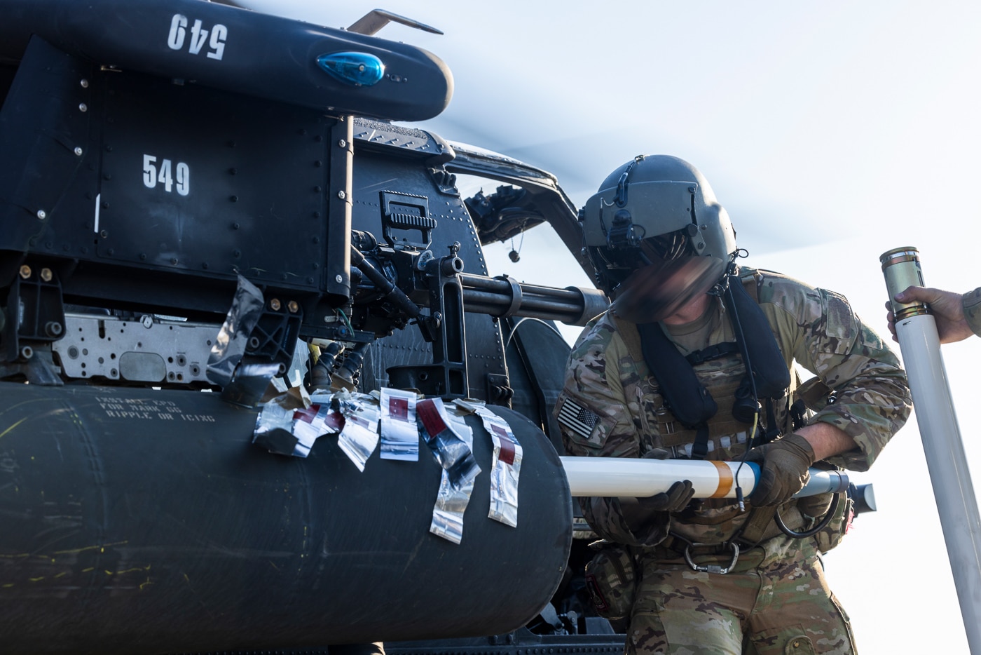 The image shows a U.S. Army Soldier assigned to the 160th Special Operations Aviation Regiment (Airborne) loading rockets into a weapons pod on the side of an MH-60 Black Hawk helicopter. The setting is Marine Corps Outlying Field Atlantic, where a forward arming and refueling point (FARP) exercise is taking place on May 6, 2025. The helicopter is parked on a field or tarmac, with its side doors open and ground support equipment visible nearby. The soldier is wearing standard flightline safety gear, including hearing protection and gloves, while carefully securing rockets in preparation for the next sortie. The FARP setup includes fuel trucks, ammunition handling equipment, and other Night Stalkers personnel working in the background. This image highlights the importance of rapid rearming and refueling operations for special operations aviation units. The scenario demonstrates the logistics and teamwork required to keep helicopters combat-ready in remote or austere locations.