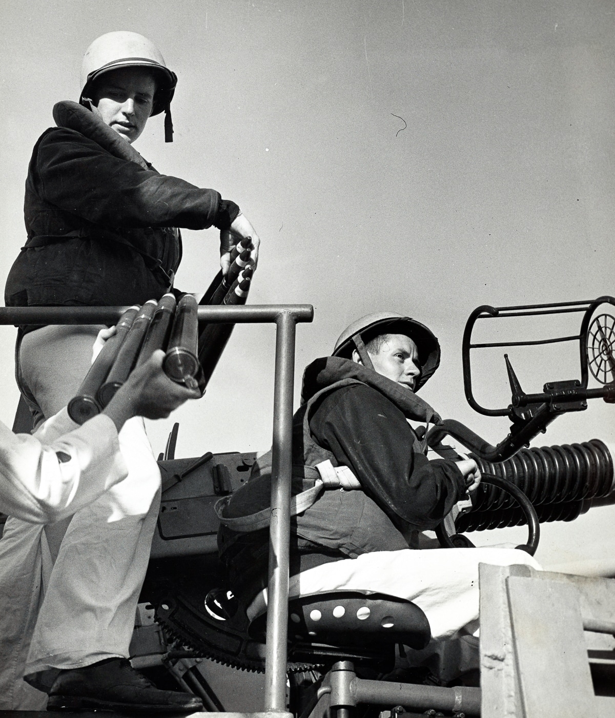 A wartime photo captures U.S. Coast Guardsmen loading 4-round aluminum ammunition clips into a 40mm Bofors anti-aircraft gun during a Japanese kamikaze attack in World War II. The gun crew works rapidly — one sailor inserts a clip while another readies the next, and the gunner maintains aim on an unseen aircraft. Spent shell casings scatter around their feet, glinting in the sunlight. The ocean horizon is visible in the background with faint smoke from anti-aircraft bursts in the distance. Each man wears a steel helmet and life vest, showing the tension of battle conditions. The image highlights the critical role of Coast Guard gunners in protecting ships from Japanese Kamikazes in World War II.