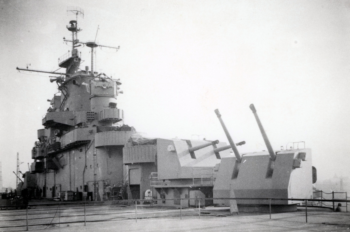 A World War II-era photo showing a 5"/38 caliber Mark 12 dual-purpose gun mount on the starboard side of an Essex-class aircraft carrier. The large twin-barrel turret is elevated toward the sky, positioned to engage approaching aircraft. Nearby, the Mark 37 Fire Control System director—an enclosed, radar-guided targeting tower—can be seen atop the carrier’s island structure. Several sailors stand by the gun mount for loading and maintenance duties. The combination of radar tracking and automatic fire control allowed the system to target kamikaze planes with high precision. This photo demonstrates the technological sophistication of U.S. Navy defenses against Japanese Kamikazes in World War II.