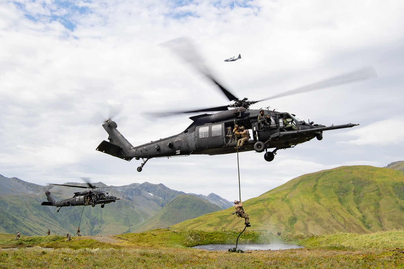 The photo shows US Navy SEALs fast-roping from an MH-60M Black Hawk helicopter operated by the 160th Special Operations Aviation Regiment (Airborne) during a training mission on Attu Island, Alaska. The helicopter hovers above the rugged, windswept terrain as SEAL operators, wearing cold-weather gear and helmets, descend rapidly on thick ropes to the ground below. The scene highlights the remote, challenging environment of Attu Island, with sparse vegetation, rocky ground, and possibly snow or mist in the background. The MH-60M is clearly marked with special operations features, including external fuel tanks and open doors for rapid deployment. The fast-rope technique allows for quick insertion of troops when landing is not possible or practical. The collaboration between the 160th SOAR and Navy SEALs is evident, showcasing joint training and operational integration. This image illustrates the adaptability and readiness of special operations aviation in Arctic and remote settings.