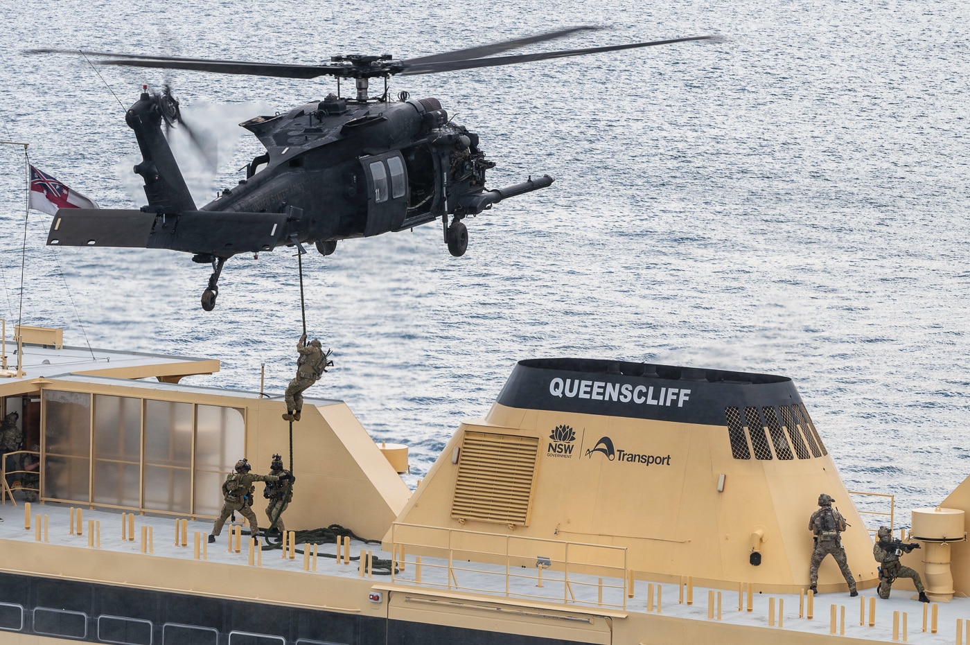 The image shows U.S. Naval Special Warfare operators, Australian Special Operations Command soldiers, and United Kingdom Royal Marine Commandos from 42 Commando conducting a Visit, Board, Search, and Seizure (VBSS) exercise on a simulated target vessel in New South Wales, Australia. A U.S. Army MH-60 helicopter from the 160th Special Operations Aviation Regiment (Airborne) hovers overhead or is parked nearby, supporting the operation. The multinational special operations teams are equipped with tactical gear and helmets, climbing ladders or ropes to board the ship, while the helicopter provides aerial security or insertion capabilities. The exercise, part of Talisman Sabre 25, brings together allied special operations forces to practice coordinated maritime boarding, search, and seizure procedures. The photo highlights the strong partnership between the U.S., Australian, and British militaries. The vessel is marked and equipped for training purposes, with crew members or role players possibly visible. This image demonstrates the operational flexibility and international collaboration central to modern special operations.
