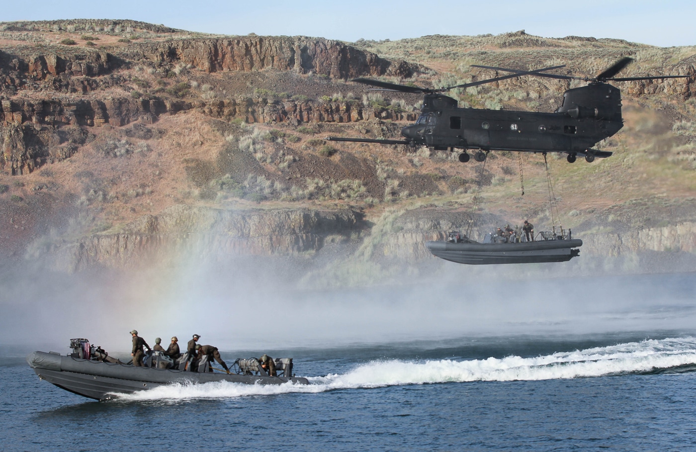 The image shows US Navy Special Warfare Combatant-craft Crewmen (SWCC) deploying from a 160th Special Operations Aviation Regiment (Airborne) Chinook helicopter during a training or operational scenario. The Chinook, with rear ramp or side doors open, hovers above water or a shoreline, allowing SWCC operators in tactical gear to exit quickly. Some crewmen may be descending ropes, ladders, or jumping into the water, prepared for immediate insertion into a maritime environment. The background may include visible watercraft or support boats, highlighting the integration of air and sea assets. The helicopter is equipped with night-vision gear and other modifications for special operations. This scene underscores the joint nature of SOAR and Navy missions, with an emphasis on rapid deployment and maritime readiness. The image illustrates the advanced training and interoperability essential for successful special operations.