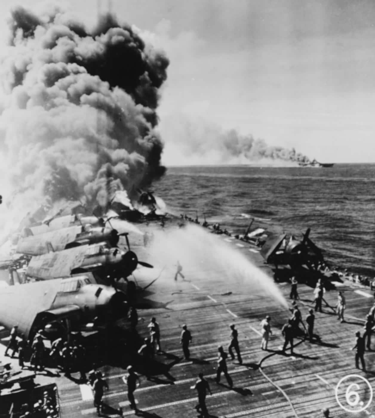 A wartime photograph showing the USS Belleau Wood (CVL-24) engulfed in flames after a kamikaze plane struck her aft flight deck during operations near the Philippines in late 1944. Thick, black smoke billows from the carrier, rising thousands of feet into the sky. In the distance, the larger fleet carrier USS Franklin (CV-13) also burns, its flight deck covered in smoke from secondary explosions. The sea between the two ships is littered with debris and oil slicks. Both vessels were part of the same task force hit by multiple suicide planes in quick succession. The image vividly conveys the devastation caused by Japanese Kamikazes in World War II and the peril faced by American carrier crews.