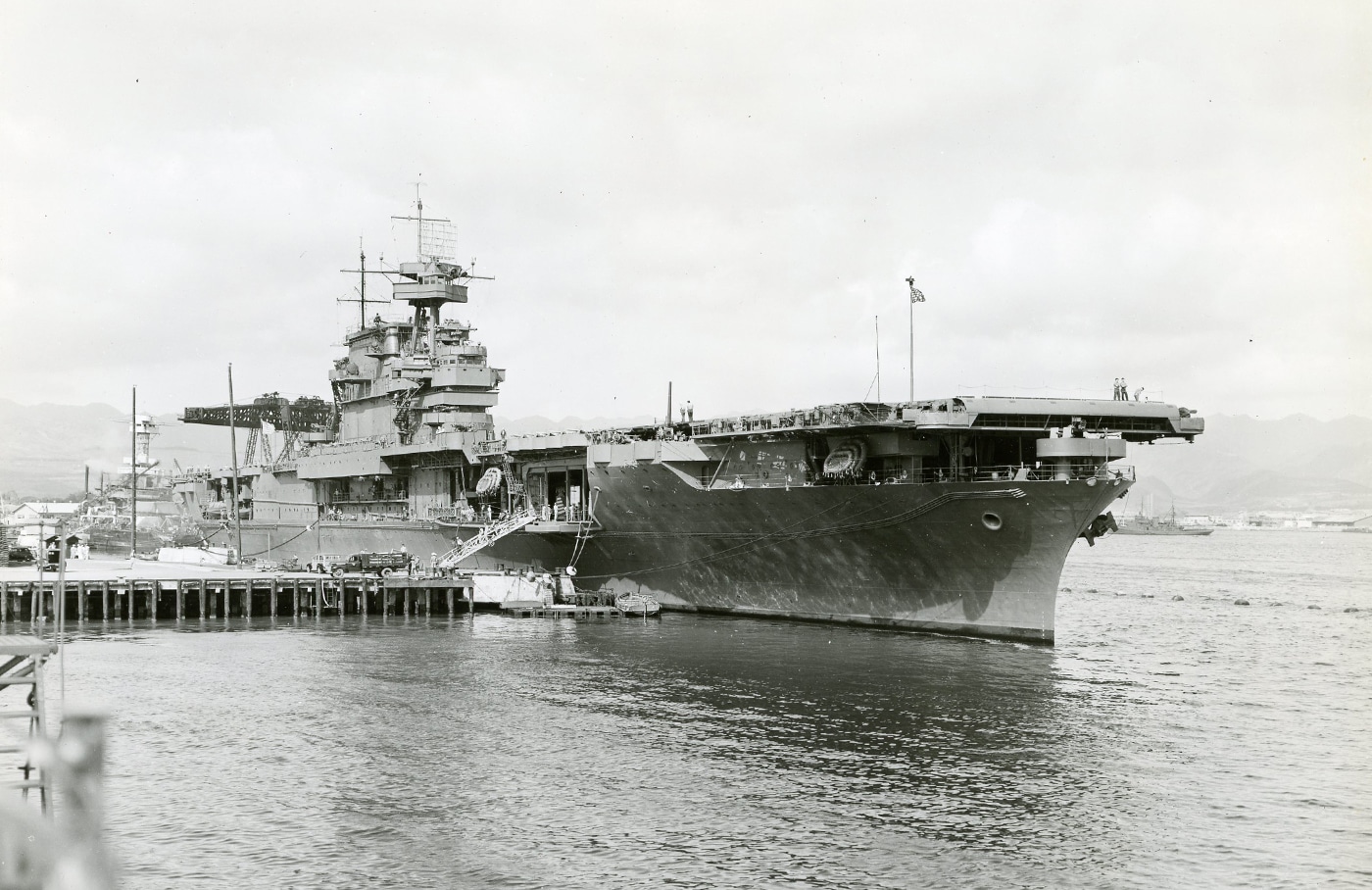 This photo shows the USS Enterprise (CV-6) docked at a port facility during World War II as supplies and munitions are being loaded aboard. The carrier’s flight deck and superstructure are visible, with cranes and cargo nets actively lifting boxes, barrels, and artillery shells onto the ship. Dock workers and naval personnel work together, moving materials and overseeing the transfer of ammunition. Trucks and supply vehicles can be seen nearby, indicating a busy and well-coordinated logistics operation. Crew members on deck are directing the process and securing loads for storage. This scene highlights the critical behind-the-scenes effort required to keep a frontline aircraft carrier supplied and combat ready. Without regular resupply, extended operations at sea would have been impossible for ships like USS Enterprise. The photo offers a detailed view of wartime naval logistics in action.