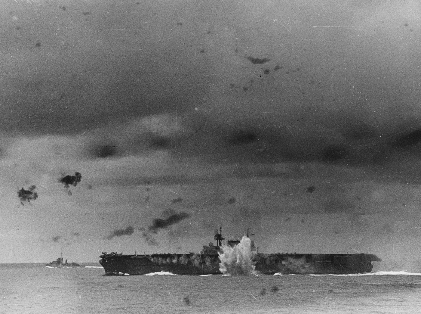 This photo captures the USS Enterprise (CV-6) during an air raid by Japanese forces off the Santa Cruz Islands in October 1942. Explosions and smoke are visible on and around the ship as Japanese aircraft execute bombing and strafing runs. Crew members can be seen at battle stations, responding with anti-aircraft fire to defend the carrier. The image conveys the chaos and danger faced during these intense attacks. Despite sustaining serious damage to her flight deck and superstructure, USS Enterprise remained afloat and operational. This event was one of several times during World War II when the carrier was reported sunk by the enemy, but she always returned to service. The carrier’s survival here was crucial to maintaining U.S. naval strength in the Pacific.