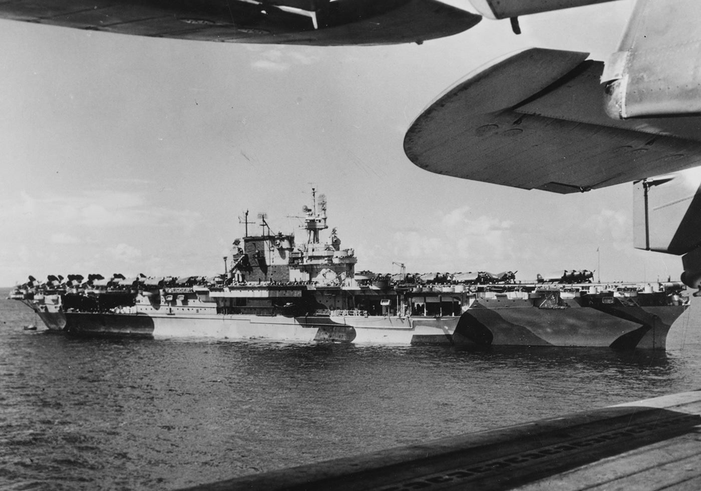 This photo shows USS Enterprise (CV-6) anchored off the coast of Saipan during World War II, her hull and superstructure painted in the distinctive Measure 33, Design 4ab camouflage pattern. The geometric blocks of light and dark gray, blue, and black create a disruptive effect, making it harder for enemy forces to estimate range and speed. The ship is stationary in calm waters, with her flight deck mostly clear and the island structure visible. Smaller support vessels and landing craft may be seen nearby, indicating ongoing operations in the area. The photo captures a key moment after the Battle of Saipan, with Enterprise serving as a mobile airbase for continued operations in the Mariana Islands. The camouflage is sharp and freshly applied, showing the Navy’s commitment to innovative defensive tactics. The image provides a rare look at one of the most iconic carriers of the war in full Pacific theater paint.