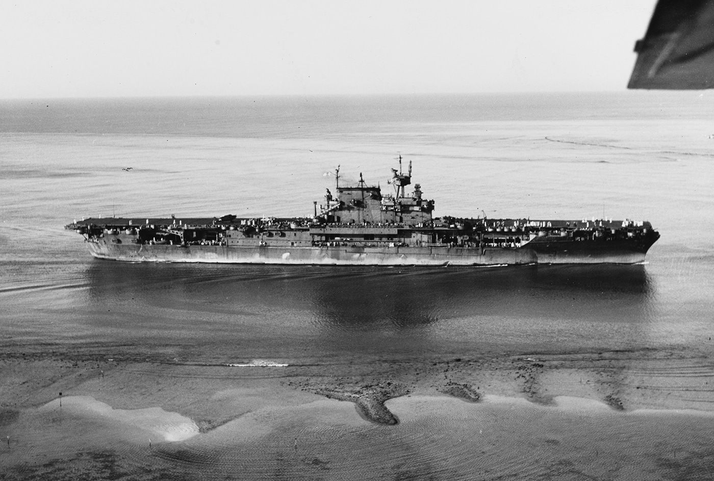 This photo shows USS Enterprise (CV-6) entering Pearl Harbor in 1943 after completing the Marshall Islands campaign. The carrier is moving slowly through the harbor channel, with crew members lined along the flight deck and superstructure, many waving or standing at attention. The ship’s hull bears signs of recent operations, and aircraft are parked neatly on deck, some with visible mission markings. Pearl Harbor’s docks, buildings, and other Navy vessels are visible in the background, providing context and scale. The mood appears to be one of relief and accomplishment as the famous carrier returns from another successful Pacific operation. The scene highlights the significance of Pearl Harbor as a major U.S. naval base and staging area during World War II. The arrival of Enterprise was often a morale boost for all personnel stationed there.