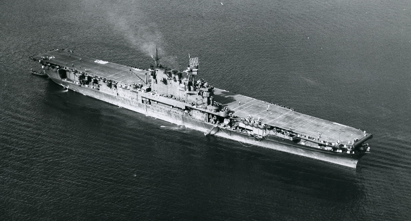 This photo shows USS Enterprise (CV-6) underway at sea in 1942, captured either from an escort ship or an aircraft flying nearby. The carrier’s flight deck is clear, with some aircraft visible, and the island superstructure rises above the deck midship. The ocean is relatively calm, allowing the ship to travel at speed, with a prominent wake trailing behind. The ship’s hull and upperworks are painted in a wartime camouflage scheme, designed to break up her outline against the water. This year was pivotal for Enterprise, as she participated in major operations including the Doolittle Raid, the Battle of Midway, and the Guadalcanal campaign. Crew are visible on the flight deck and in observation posts, indicating a high state of readiness. The photo gives a strong sense of the scale and power of this historic Yorktown-class carrier during one of the most intense periods of World War II.
