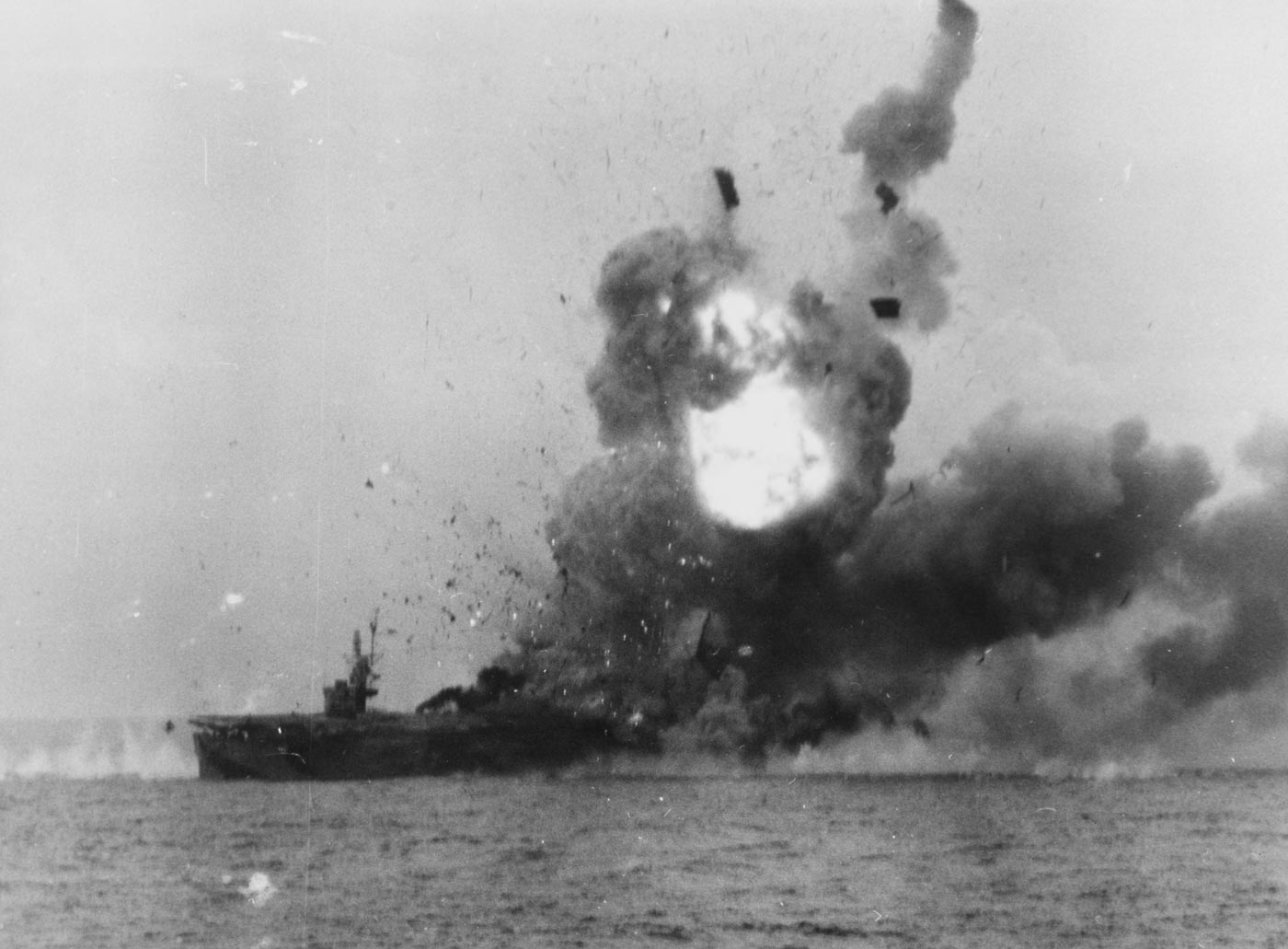 A wartime photograph shows the escort carrier USS St. Lo (CVE-63) exploding violently after being struck by a Japanese kamikaze aircraft during the Battle of Leyte Gulf on October 25, 1944. A massive column of fire and smoke rises hundreds of feet above the ship, completely obscuring her flight deck. Debris and burning fragments rain down into the sea, where oil slicks and wreckage float nearby. The carrier’s island and gun positions are barely visible through the dense black smoke. The kamikaze impact detonated bombs and aviation fuel, triggering a catastrophic secondary explosion. This image captures one of the most famous moments of Japanese Kamikazes in World War II — the destruction of St. Lo, the first U.S. carrier lost to a suicide attack.