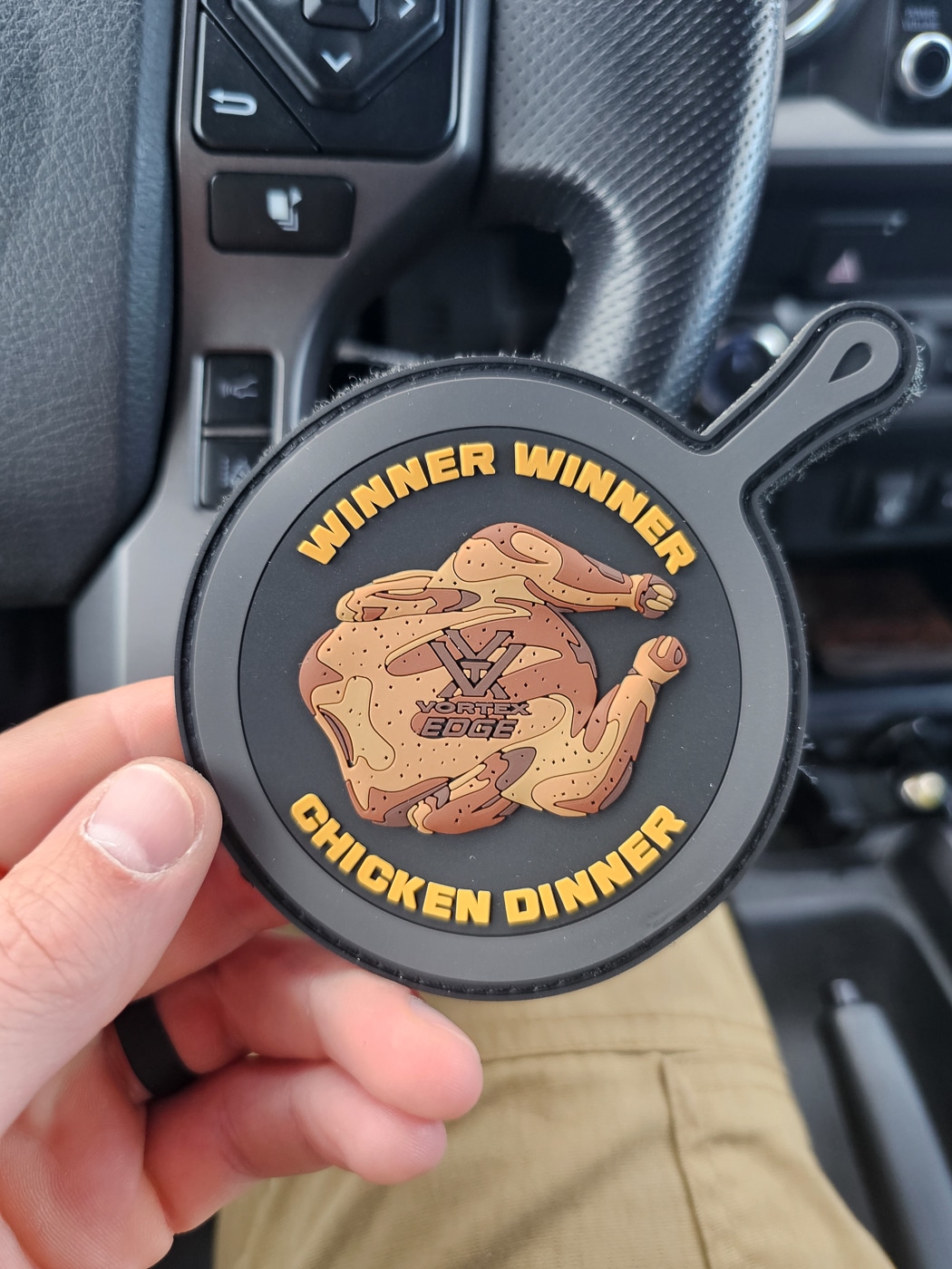 A close-up photo of the Vortex Optics “Winner Winner Chicken Dinner” morale patch awarded to the top shooter in the Hunter Marksmanship Course. The patch features a stylized chicken graphic with bold text on a circular fabric background, edged in stitching for durability. It’s laid flat on a table next to rifle accessories, ammunition boxes, and other course memorabilia. The patch serves as a playful recognition of top performance during the Field Course Test, blending competition with camaraderie. The photo captures the tone of the course  —  serious training, but with a sense of fun and community among shooters. It symbolizes proficiency, humility, and mastery of fundamentals under realistic hunting conditions.
