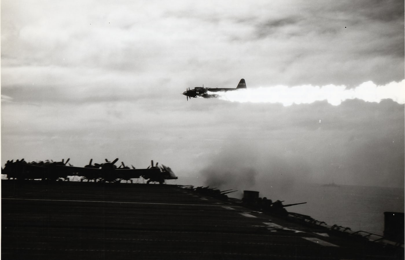 Black-and-white wartime photograph showing a Yokosuka P1Y “Frances” twin-engine plane in a low, fast descent toward the escort carrier USS Ommaney Bay (CVE-79) during a kamikaze attack. The carrier’s flight deck is visible in the foreground with silhouetted sailors and parked aircraft. Anti-aircraft bursts and splashes appear between the camera and the plane, indicating heavy defensive fire. Smoke trails from the P1Y’s engines are visible, suggesting damage or full-power approach. This image documents a suicide plane attack during the Marianas/Philippine operations and is directly related to Japanese Kamikazes in World War II. The scene highlights the vulnerability of escort carriers to kamikaze tactics and the frantic defensive efforts of ship crews. Overall, the photo conveys speed, danger, and the small-window timing defenders had to stop a kamikaze strike.