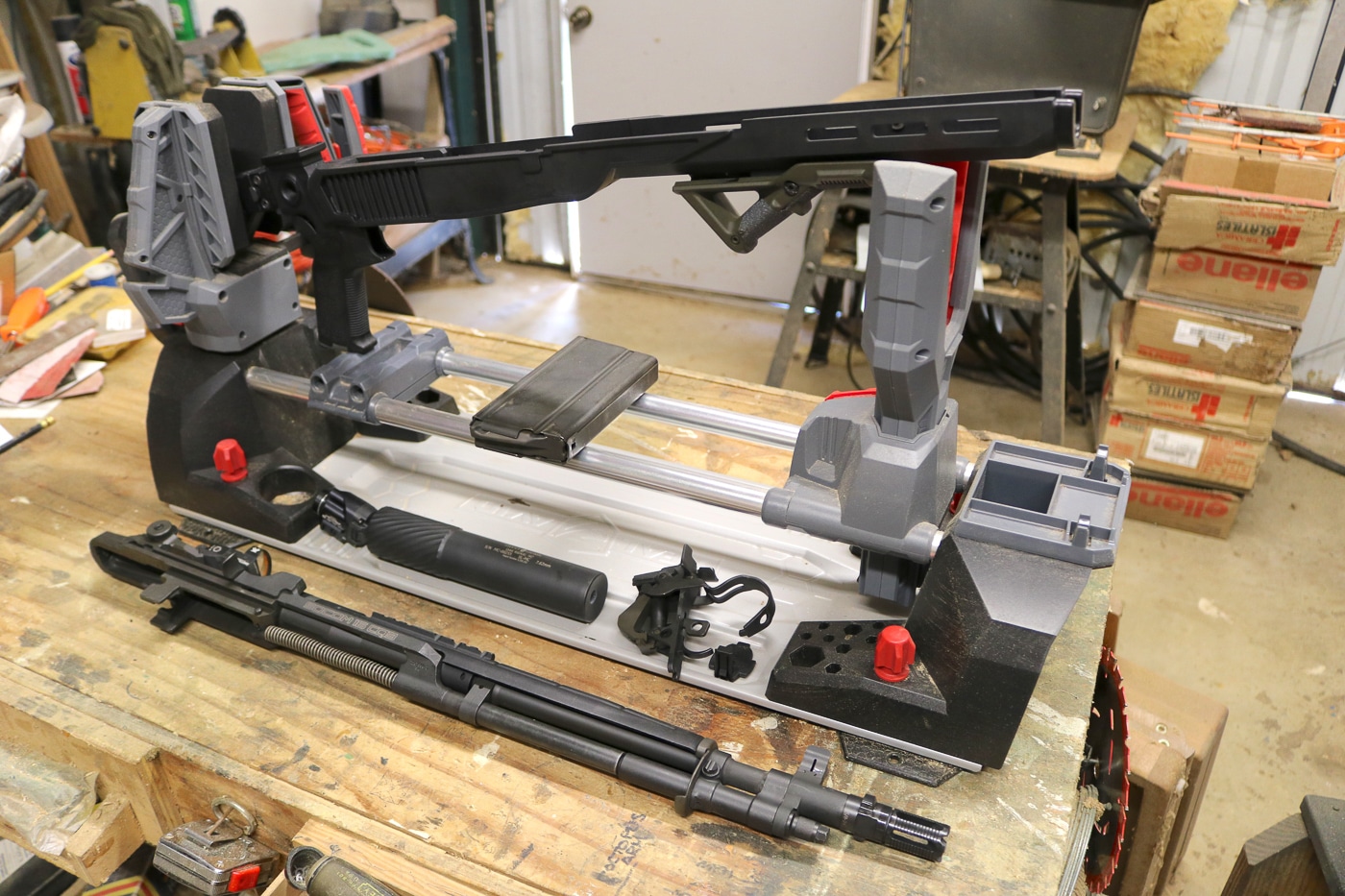 Disassembled Springfield Armory M1A SOCOM rifle and Delta 14 Chassis Gen 2 components arranged in workshop setting showing all parts required for installation. Visible are the M1A barreled action with gas system, trigger group assembly, Delta 14 glass-filled polymer chassis, and Chisel Machining aluminum folding buttstock. The layout demonstrates the drop-in nature of the chassis system requiring only separation of the trigger assembly from the action followed by placement into the new chassis. No permanent modifications to the firearm are necessary and installation can be completed by anyone capable of basic field stripping. The lightweight 0.75-pound polymer chassis features M-Lok slots, flared magwell, steel front ferrule, and Mossberg 500 pattern rear mount. Chisel buttstock includes adjustable KICK-EEZ cheekrest, adjustable recoil pad, and AR-pattern Magpul MOE-K2 pistol grip. Complete system transforms the traditional M1A into a modern tactical platform.