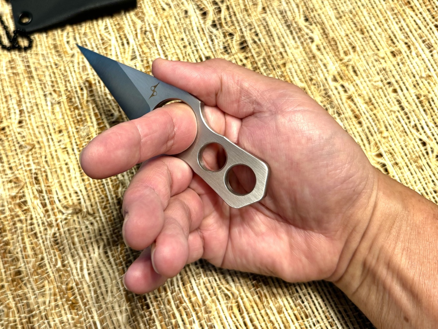 The author demonstrates holding a compact neck knife with a forward finger ring, showing how the index finger locks through the ring for retention. Only three fingers are wrapped around the short handle, but the grip remains secure and comfortable. The knife is a fixed-blade style with a slim polymer sheath visible nearby, meant for neck carry. The finger ring design prevents accidental dropping and helps with indexing under clothing. This photo emphasizes how smaller handles can still be practical when paired with a ring feature. The demonstration highlights both safety and usability for visually impaired readers, using keywords like neck knife, finger ring grip, and secure carry.