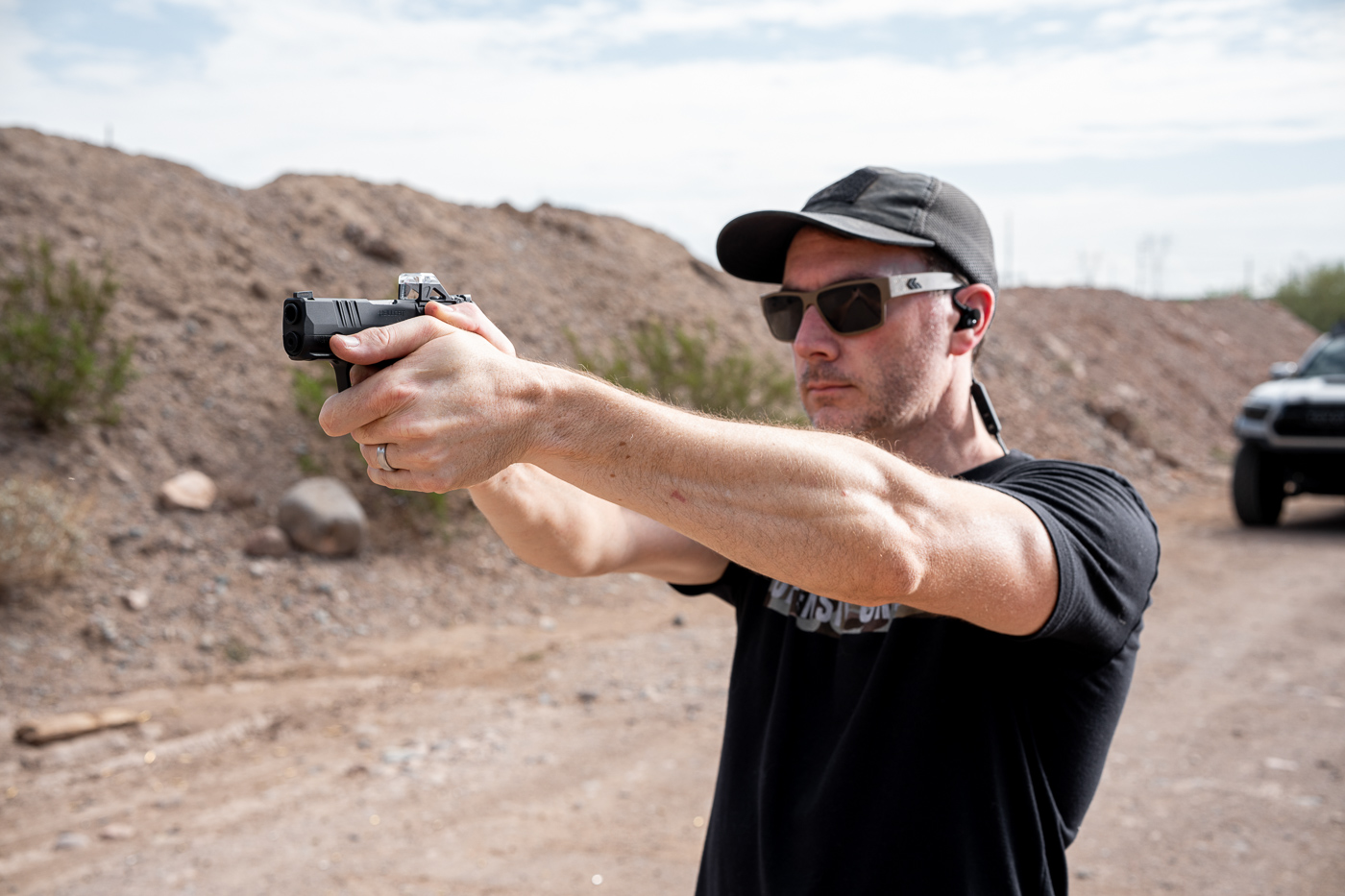 A man stands on an outdoor shooting range, firing a Springfield Hellcat 380 ACP pistol during a live-fire test. The shooter wears eye and ear protection, with arms fully extended in a two-handed grip. The pistol’s slide is mid-cycle, showing ejection of a spent casing. Dust and light in the background indicate a sunny range environment typical for firearm testing. The compact size of the Hellcat .380 is evident in the shooter’s hands, reinforcing its micro-compact classification for concealed carry. This image captures the review author testing accuracy, recoil, and real-world performance. The photo communicates hands-on evaluation and field conditions for the Springfield Armory Hellcat 380 ACP pistol.