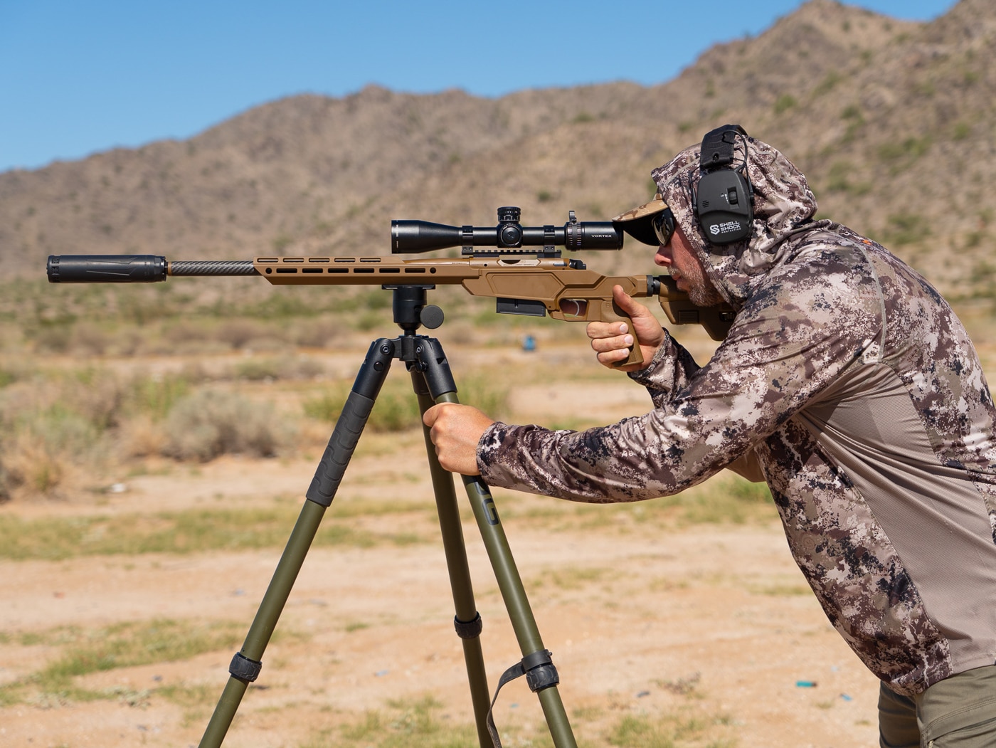 Shooter positioned behind Springfield Model 2020 Heatseeker precision rifle mounted on BOG Deathgrip tripod during range session in Arizona desert terrain. The bolt action chassis rifle with Coyote Brown Cerakote finish sits securely in tripod clamp system allowing for stable shooting platform at various angles. Vortex Viper scope mounted on rifle provides magnified view of distant targets while shooter maintains proper cheek weld on adjustable B5 Systems stock. Blackout Defense Acra plate attached to M-Lok rail provides tripod mounting point for the lightweight aluminum chassis. This shooting position allows engagement of steel plates at extended ranges beyond typical bipod capabilities. Tripod setup particularly useful for positional shooting and longer distance target work where stability matters most.