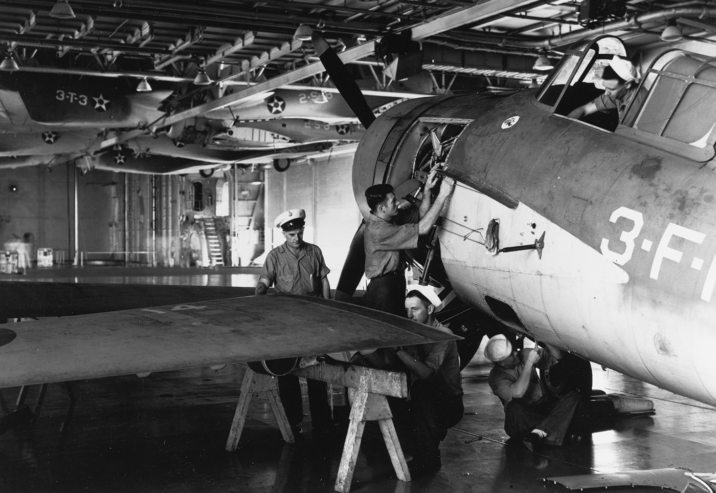 This photo shows a team of aviation machinists aboard USS Enterprise (CV-6) working on a Grumman F4F Wildcat fighter inside the carrier’s hangar deck. The mechanics are gathered around the aircraft, with engine panels open and various tools in use as they perform routine maintenance or repairs. The hangar deck is busy, with other crew members moving in the background and parts or equipment neatly organized along the bulkheads. Lighting is provided by overhead fixtures, illuminating the details of the aircraft and the machinists’ focused efforts. The F4F’s distinctive folding wings and U.S. Navy markings are visible. Regular maintenance like this kept Enterprise’s air group flying constant missions in the Pacific. The image highlights the technical expertise and teamwork required to keep complex machinery combat-ready at sea. It’s a detailed look at the behind-the-scenes work critical to the carrier’s success.