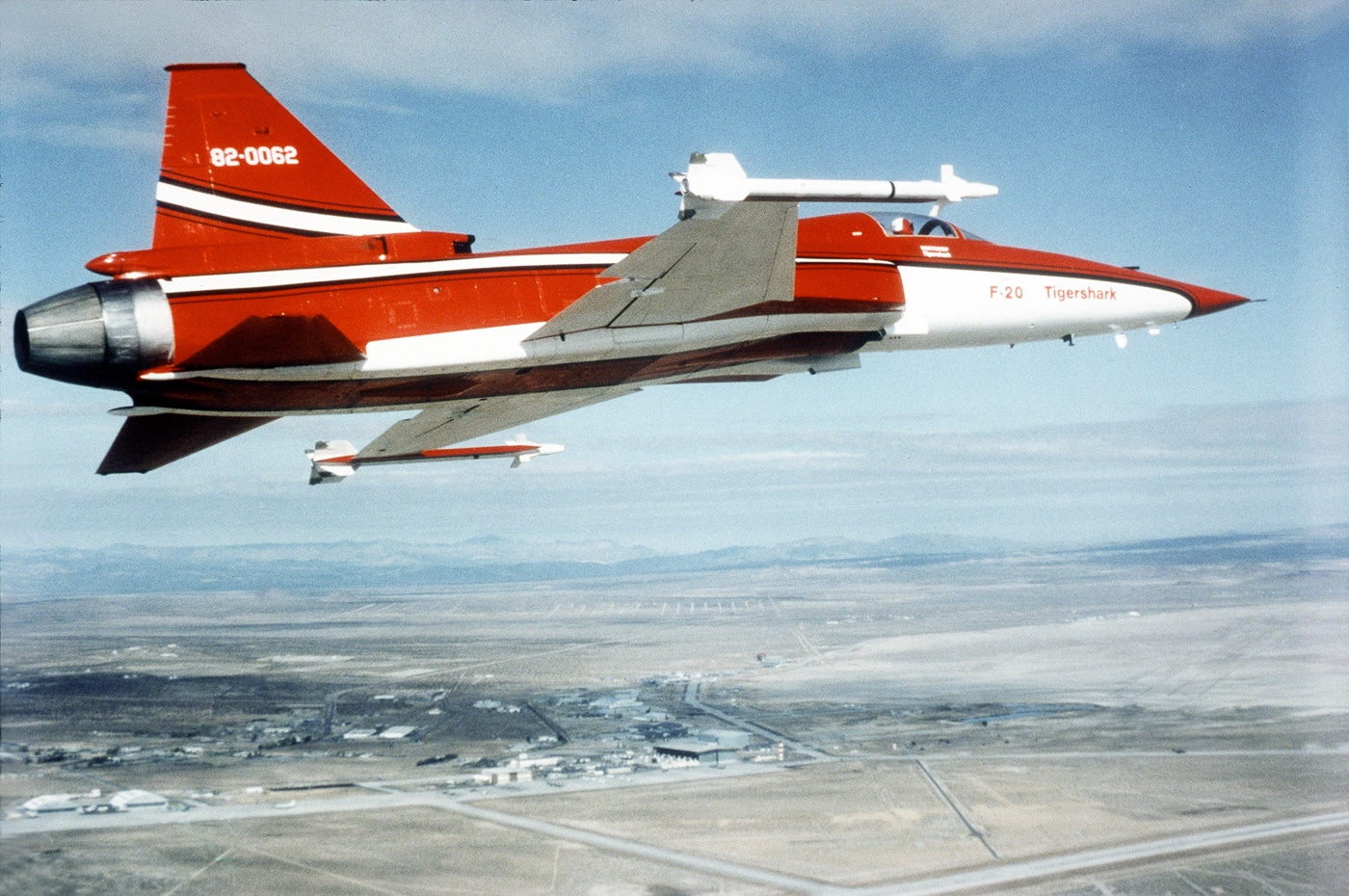 A right-side, air-to-air photograph shows a Northrop F-20 Tigershark flying in a red and white “glamor” paint scheme used for early demonstrations. The aircraft is in level flight under clear skies, its polished red upper surfaces and white lower fuselage glinting in the sunlight. The F-20’s sleek design, single engine intake, and swept wings are sharply visible. The livery includes large white markings on the tail and fuselage, emphasizing its prototype identity. Northrop painted the Tigershark in this eye-catching color combination to make it recognizable at airshows and media events. The image highlights the smooth aerodynamic lines and modern features that differentiated the F-20 from the older F-5. The background sky provides a clean, unobstructed view, making the aircraft the clear focus of the composition.
