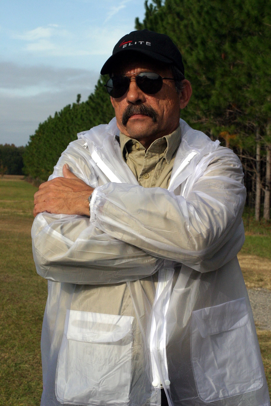 A middle-aged man stands with his arms folded across his chest, subtly resting one hand near the opening of a shoulder holster, sometimes called a Fudd holster. The shoulder rig is worn under the arm and the harness straps cross the upper chest, but the clothing keeps the setup visually low-key. His posture looks casual, as if engaged in conversation, showing how a carrier might keep a hand on a holster without drawing attention. The scene emphasizes concealment and discreet readiness rather than an active draw or demonstration of technique. Lighting is neutral and the background uncluttered so the overall impression is about situational posture and gear choice. The image ties to the article’s broader discussion of when shoulder rigs make sense for concealed carry, balancing comfort and accessibility.