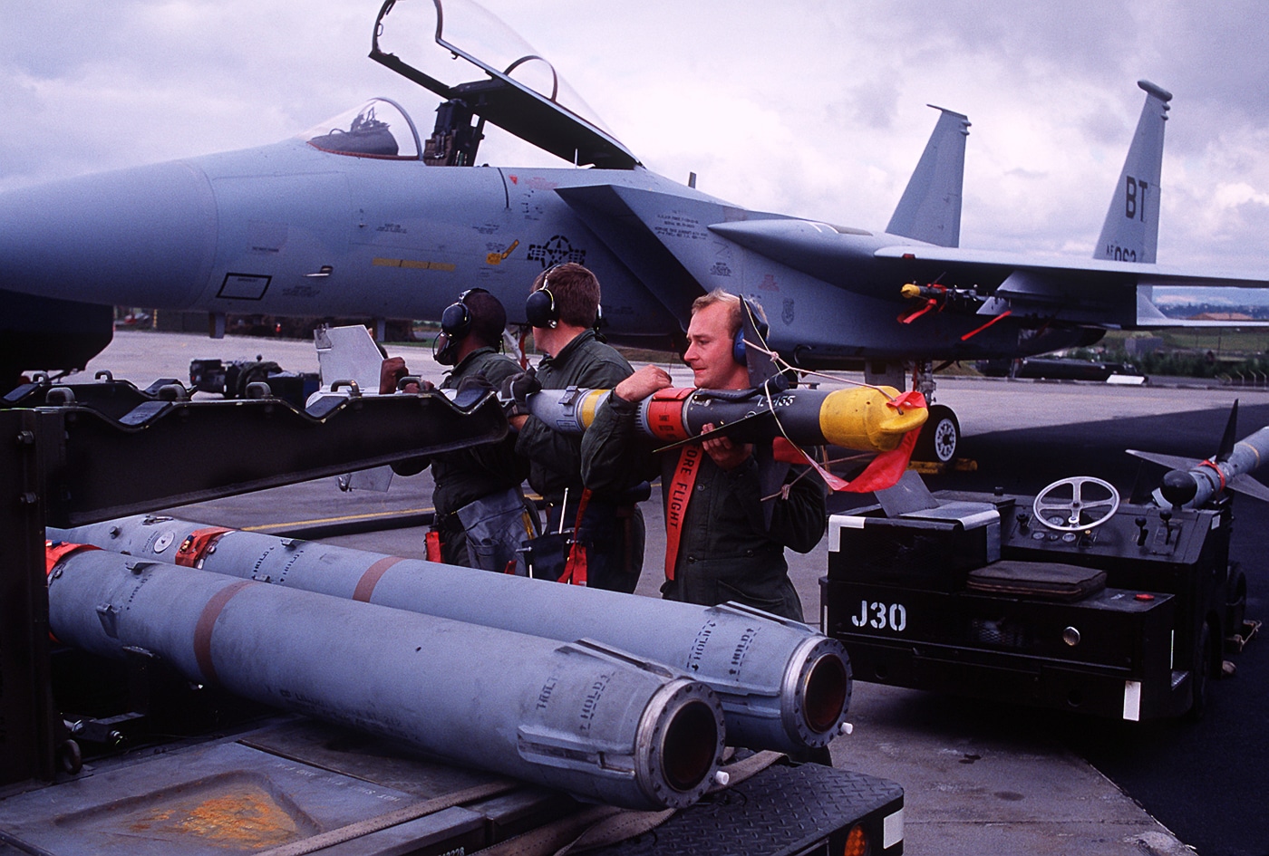 A U.S. Air Force ground crew loads an AIM-9 Sidewinder missile onto an F-15C Eagle fighter at Bitburg Air Base, West Germany. The crew members work beneath the aircraft’s wing, guiding the missile onto its pylon mount with specialized equipment. The F-15C’s distinctive twin tails and gray camouflage paint scheme are visible in the background. This photo reflects standard weapons loading procedures that ensured aircraft remained ready for immediate scramble during the Cold War. The AIM-9 Sidewinder, a short-range infrared-guided missile, was a primary air-to-air weapon used by NATO forces. Bitburg Air Base served as a major hub for U.S. Air Force operations in Europe throughout the 1980s. The image captures the balance of precision, safety, and speed required in front-line aircraft maintenance.