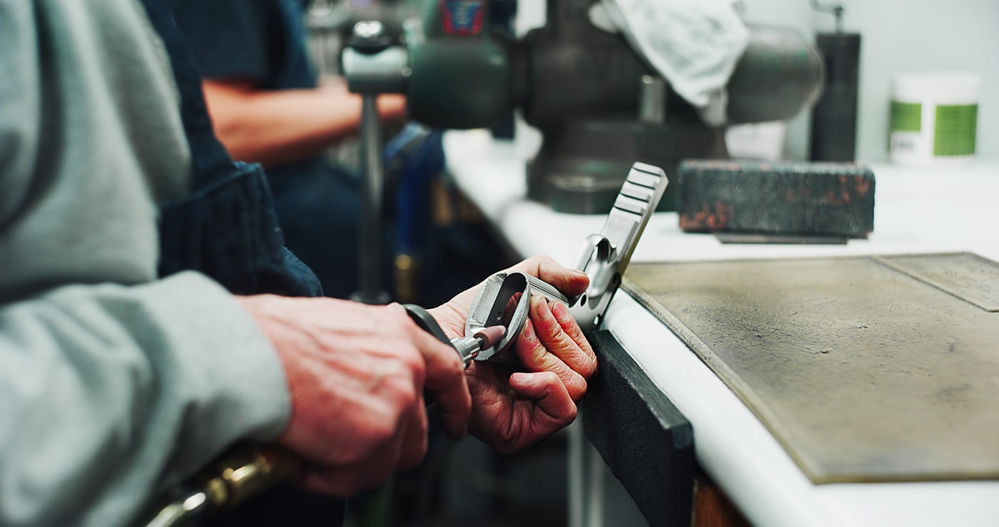 Professional gunsmith performing custom magazine well work on a 1911 pistol frame to improve reload speed and ease of use. The beveling process requires specialized tools and expertise that significantly increases the final cost of custom builds. Plain jane 1911 models function reliably for defensive purposes without enhanced magazine wells or competition-grade modifications. Custom shop services can add thousands of dollars to basic pistol prices, with top-tier guns costing $3,000 or more. Standard configuration models like the Springfield Armory Mil-Spec at $725 feed dependably without beveled mag wells. The practical benefit of faster reloads matters more for competition shooting than real-world defensive encounters. Gunsmiths have offered magazine well improvements since the 1960s, but military and law enforcement used unmodified 1911 pistols effectively for decades.