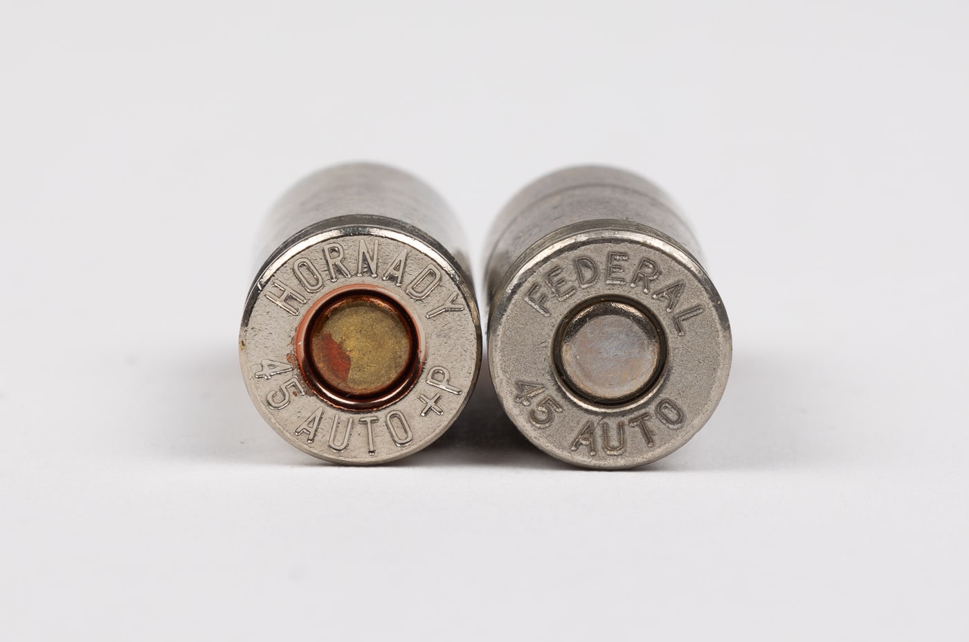 Photograph featuring two ammunition cartridges showing headstamp markings with one displaying plus P overpressure designation and one showing standard pressure specifications. Cartridge headstamps contain crucial safety information about ammunition pressure levels. Plus P ammunition can be identified by specific markings stamped on the cartridge base. Standard pressure loads show different headstamp information without overpressure ratings. Firearm safety requires matching ammunition pressure specifications to gun manufacturer ratings. The headstamp comparison illustrates differences between higher pressure plus P loads and regular pressure ammunition. Shooters must verify cartridge markings before using overpressure ammunition in any firearm. Understanding these stamped designations prevents using plus P rounds in guns not designed for increased pressure loads.