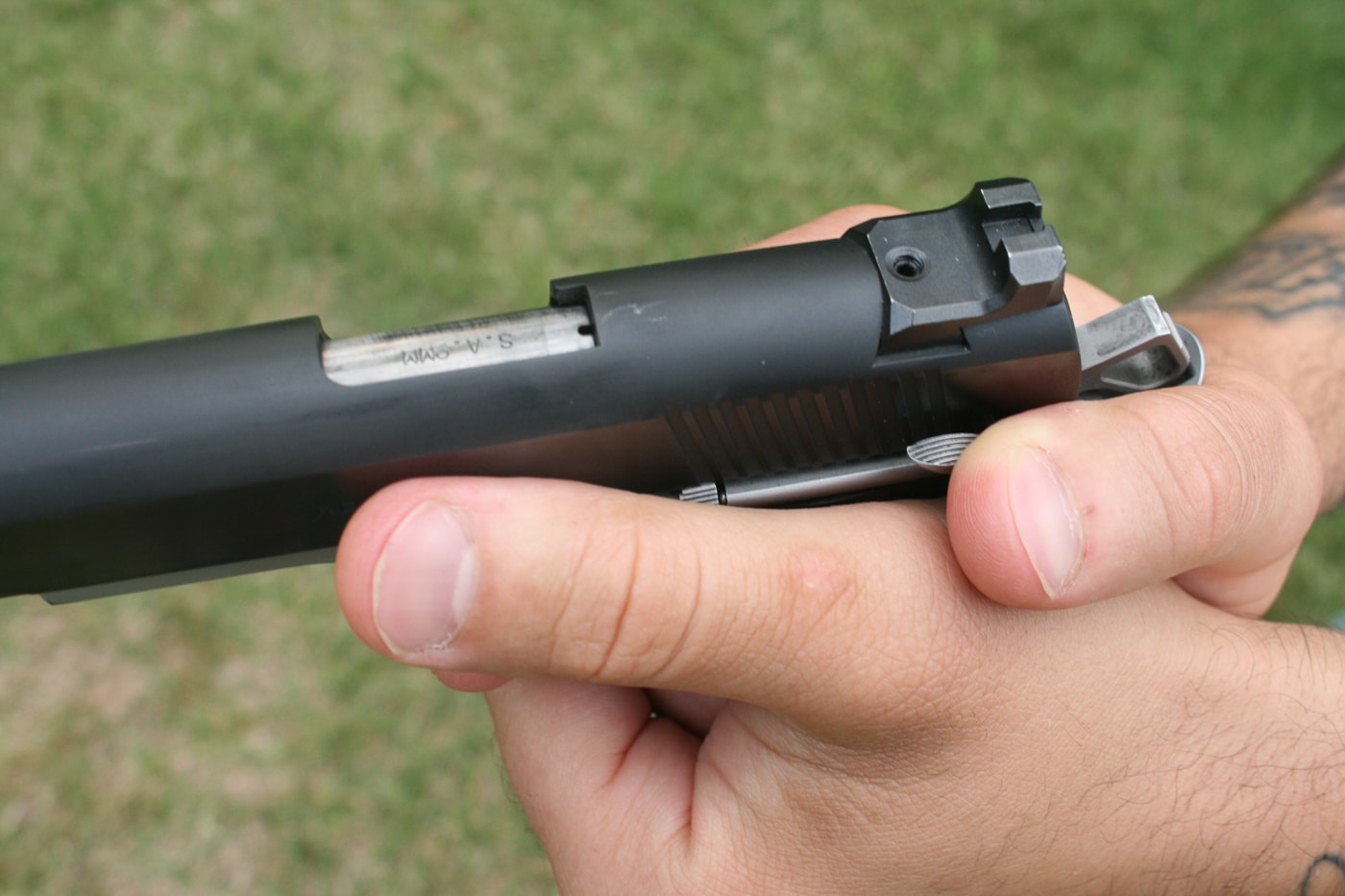 Close-up image demonstrating thumbs-forward grip fundamentals on a 1911-style pistol showing proper hand indexing and backstrap contact. The shooter's dominant hand thumb maintains contact with the frame-mounted safety allowing instantaneous manipulation to sweep the safety down into action or back up to place the pistol on safe. Middle finger knuckle of the dominant hand is in hard contact with the underside of the trigger guard to mitigate muzzle dip when shooting at speed. Support hand is placed with palm contact on the grip panel creating a firm two-hand connection to the pistol. Pay attention to your grip when shooting because this high grip position brings the long bones of the arm as close as possible to the axis of the bore minimizing muzzle flip. All fingers tightly encircle the grip frame providing the firm pressure needed for proper trigger control and sight alignment.