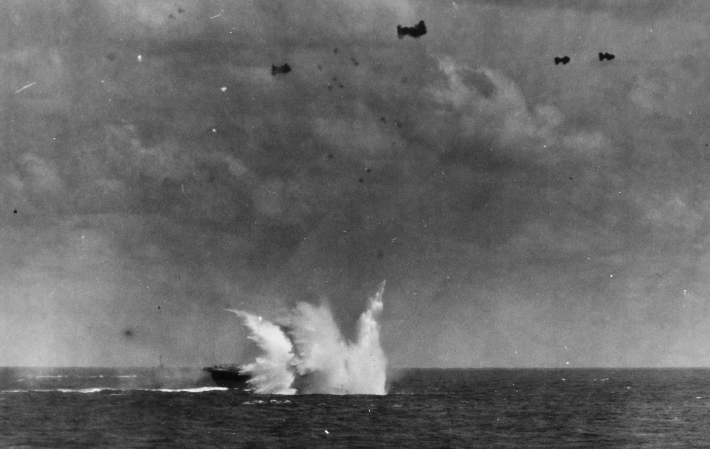 This dramatic photo shows a Japanese kamikaze aircraft hitting the water next to USS Enterprise (CV-6) during combat operations off Okinawa in April 1945. The aircraft impacts the sea just yards from the carrier, sending a massive splash and spray into the air alongside the ship’s hull. The carrier’s flight deck, with aircraft and crew members visible, is shown at action stations, ready for immediate response. The event demonstrates how close the kamikaze threat came to inflicting serious damage or sinking major Allied warships. Enterprise’s anti-aircraft guns and evasive maneuvering likely contributed to the near miss. The background reveals the expanse of the Pacific and the urgency of ongoing combat. Kamikaze attacks were a frequent and deadly danger in this phase of the war. The image captures both the luck and the constant tension of carrier life at Okinawa.