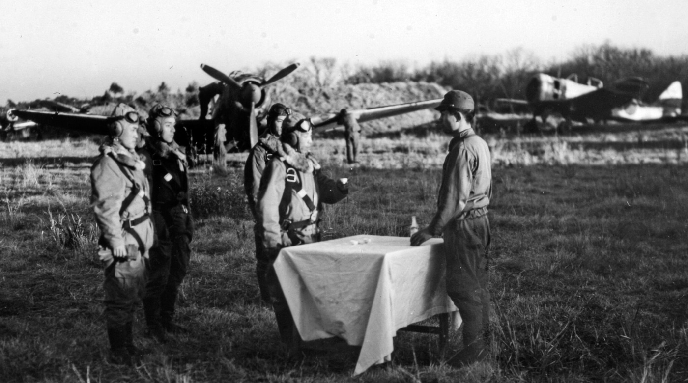 A historical photo showing a group of young Japanese kamikaze pilots standing beside their aircraft before a mission during World War II. They wear flight suits, life vests, and hachimaki headbands with patriotic symbols. Ground crew members and officers can be seen in the background offering final words and salutes. The mood is solemn, reflecting the pilots’ awareness of their one-way assignment. The planes are light, single-engine fighters lined up on an airfield under cloudy skies. This image represents the human face of Japan’s kamikaze program and the desperation of late-war defense efforts.