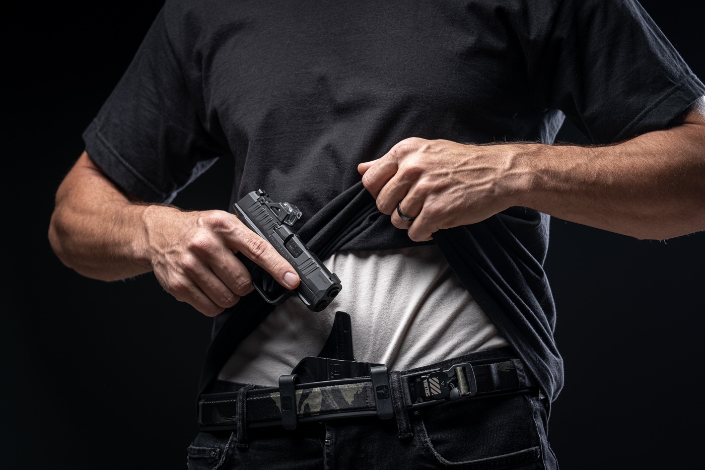 A man draws a Springfield Hellcat 380 ACP from an inside-the-waistband appendix holster; the photo shows the gun partially clear of the holster with the shooter’s strong hand on the grip. The image highlights appendix carry (AIWB) presentation with a micro-compact pistol designed for concealed carry. Visible details include the Hellcat’s textured grip, short slide and compact profile that reduce printing under clothing. The shooter’s trigger finger is held visibly outside the trigger guard, indicating safe handling during the draw. The holster is a molded Kydex-style appendix holster that keeps the pistol close to the body for concealment and speed. This photo is suitable for illustrating real-world carry, draw mechanics, and the Hellcat OSP/.380 ACP ergonomics in a review context.
