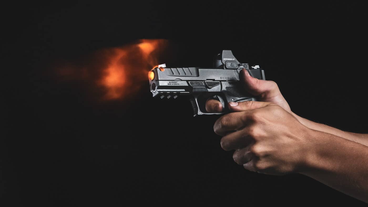 Action photograph capturing proper pistol grip technique on a Springfield Armory Echelon during shooting practice. Both hands work together with the dominant hand positioned as high as possible on the backstrap and support hand providing complementary pressure on the opposite side. Thumbs are oriented forward along the frame without interfering with external controls or slide operation. The grip demonstrates how fingers tightly encircle the grip frame while maintaining trigger finger placement on the trigger face. Establishing this type of firm two-hand grip connection to the pistol is foundational for defensive-style shooting success. Proper hand placement and grip pressure directly impact your ability to control recoil manage muzzle flip and deliver accurate shots on target.