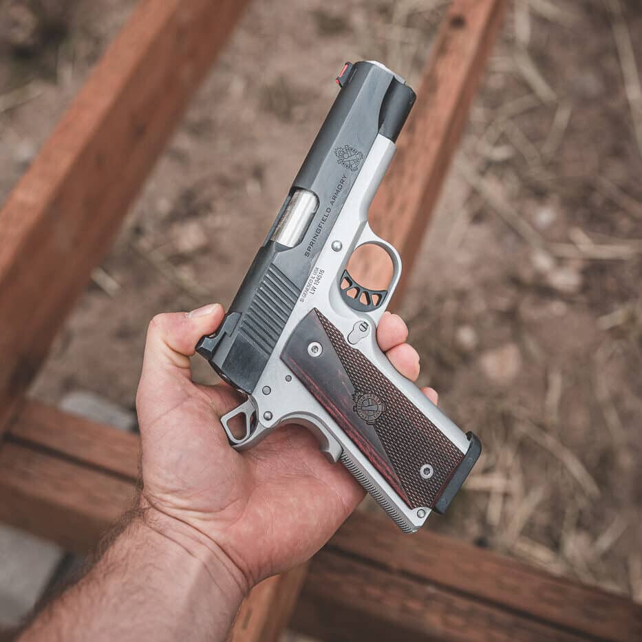 Photograph showing a shooter's hand approaching a Springfield Armory Ronin 1911 pistol before establishing final grip position. The dominant hand is positioned near the pistol demonstrating the initial contact point where proper grip begins. This image illustrates the importance of hand placement during the draw stroke and how shooters should index the web of their hand high on the backstrap. The 1911 design features a pronounced tang and recessed area that serves as a reference point for correct grip establishment. Proper initial hand positioning is a critical fundamental that affects all subsequent aspects of defensive shooting including trigger control and sight alignment. Pay attention to your grip when shooting by ensuring correct hand placement from the very first moment of contact.