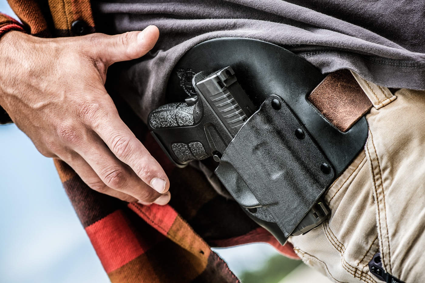 Photograph showing a shooter's hand reaching to draw a Springfield XD-S pistol from a strong side holster demonstrating the critical moment when grip establishment begins. The dominant hand is positioned to make contact with the pistol grip frame while still holstered. This initial contact point is where proper defensive shooting fundamentals start because the shooter must achieve a firm and final shooting grip before beginning the draw stroke. The holster design must allow the shooter to index the web of the hand high on the backstrap and establish proper finger placement around the grip frame. Any security devices on the holster should be releasable without compromising the shooter's ability to get a high grip on the pistol. Pay attention to your grip when shooting by recognizing that grip quality is determined from this very first moment of hand-to-pistol contact.