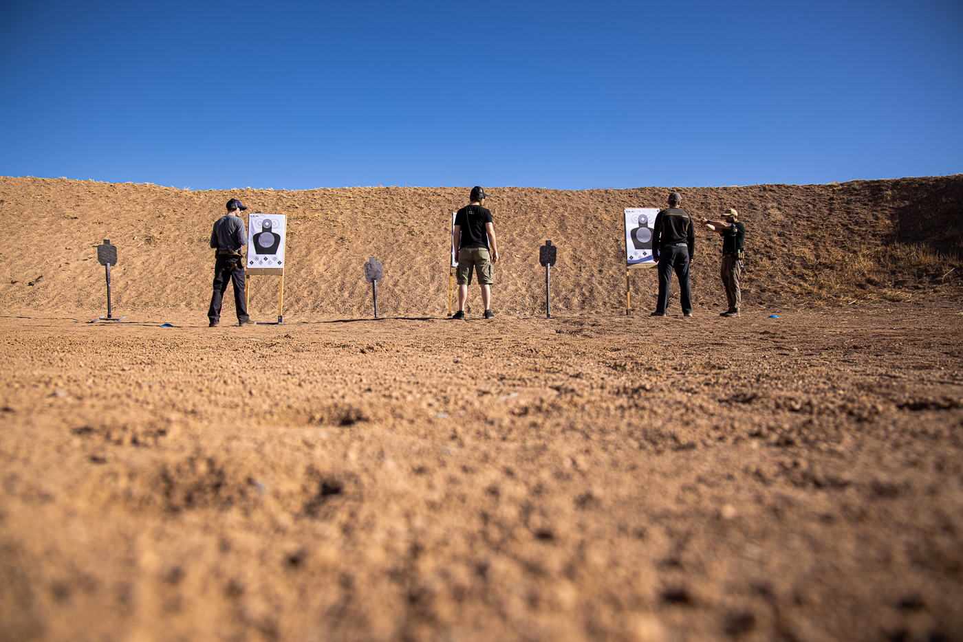 At an outdoor shooting range, a firearms instructor is giving detailed instructions to a shooter about the upcoming drill. The instructor is gesturing toward the training area, ensuring the student understands the sequence of movements. This part of the session emphasizes how important clear communication is before running tactical exercises. The environment includes target stands and open space for safe movement. The lesson relates directly to two self-defense shooting drills that focus on cover, reloading, and accurate fire under stress. The instructor’s role highlights the value of professional guidance when practicing defensive skills.