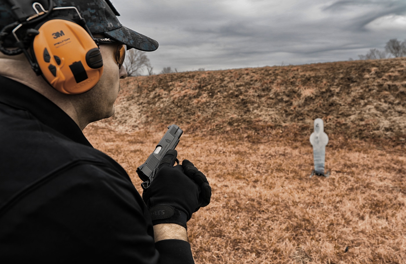 A man holding a Springfield Armory 1911 handgun stands on an outdoor shooting range preparing for training. He is dressed casually and positioned near target stands, setting up for practice. This photo represents the beginning stage of defensive firearms training where stance and grip are emphasized. The scene highlights how shooters prepare before running structured drills that simulate real-world threats. This training ties into two self-defense shooting drills that focus on movement, cover, and accuracy. The shooter is about to engage in life-saving defensive skills practice.