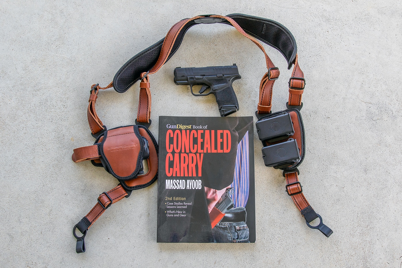 A compact modern pistol is shown secured in a shoulder holster, sometimes called a Fudd holster, positioned under the arm of the wearer. The holster harness crosses the upper chest and shoulders and holds the handgun in a horizontal under-arm position. Beside the rig sits a book about concealed carry and defensive shooting by a noted firearms author, suggesting instructional context. The scene is staged on a neutral background to focus attention on the carry system and the training material. Lighting is even and non-dramatic so details of the holster, magazine pouch, and pistol silhouette are clear. This image introduces the article’s theme of pairing traditional shoulder rigs with modern compact pistols for everyday concealed carry.