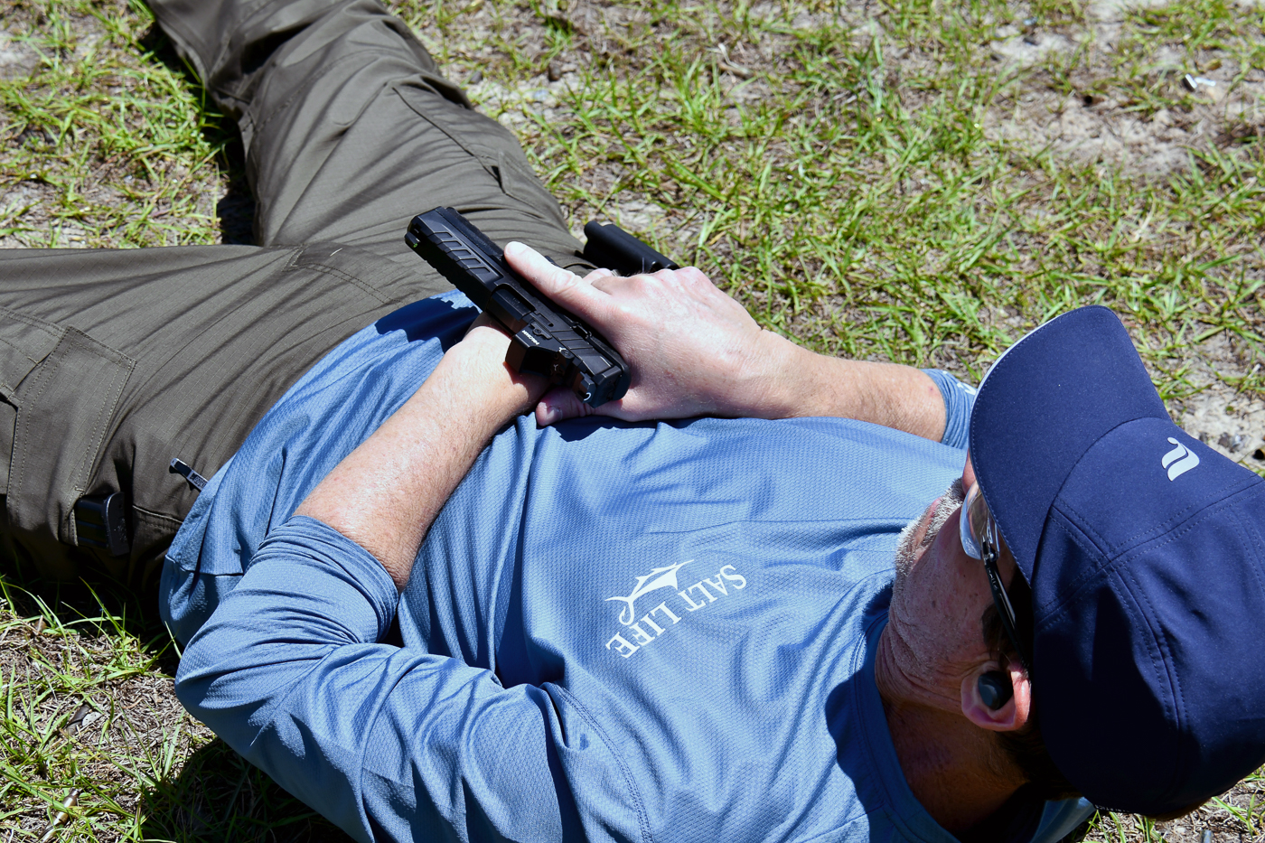 A shooter lying supine on the ground draws his pistol and starts orienting it toward the target during a training drill. His upper body is slightly lifted, and the gun is being positioned safely before firing. This stage of the exercise simulates responding to an attacker while down on the ground. Paper targets and range equipment can be seen in the background, creating a controlled environment. The drill forms part of two self-defense shooting drills designed to improve survival skills under pressure. Training like this highlights movement, positioning, and accuracy from a disadvantaged start.