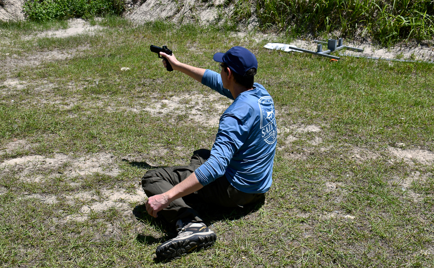 A man is shown mid-drill as he begins to stand up from the ground while keeping his handgun aimed at the targets. His posture shows deliberate, controlled movement with the muzzle pointed safely downrange. This drill simulates recovering from being knocked down during an attack and emphasizes never losing focus on the threat. The training bay includes paper targets and simulated cover, ensuring a realistic environment. This sequence is part of two self-defense shooting drills that teach shooters to handle movement under stress. The moment demonstrates how to balance safe gun handling with physical transitions.