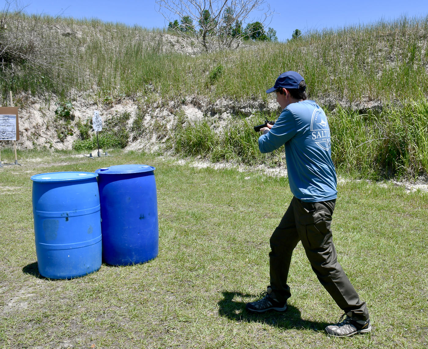 A man is captured mid-drill on an outdoor shooting range as he moves quickly toward cover while holding his handgun. His eyes remain on the threat area and his body is angled toward a plastic drum positioned as protective cover. This image illustrates how defensive training integrates movement with weapon handling. The action is part of two self-defense shooting drills that focus on using obstacles to reduce exposure while engaging targets. Paper targets and range props provide a structured environment that replicates real-life conditions. The photo emphasizes the importance of practicing not just shooting, but also movement and cover tactics.