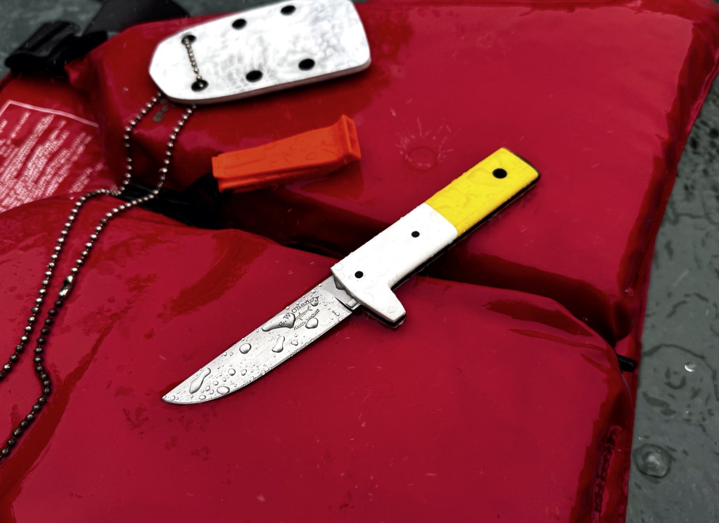A handcrafted neck knife painted in white and yellow to mimic a classic bait bucket is laid on top of boating gear including a folded life jacket and coiled dock line. The fixed-blade is housed in a molded polymer sheath and suspended on a short breakaway lanyard visible at the sheath’s throat. The color scheme is bright and high-contrast, helping the neck knife stand out when stowed among wet equipment. The photo shows water-ready styling and implies corrosion-resistant finishes suited for marine use. This descriptive view helps a visually impaired reader understand the knife’s appearance, carry method, and intended environment. Keywords: neck knife, pendant knife, boating knife, fixed-blade, sheath, lanyard.