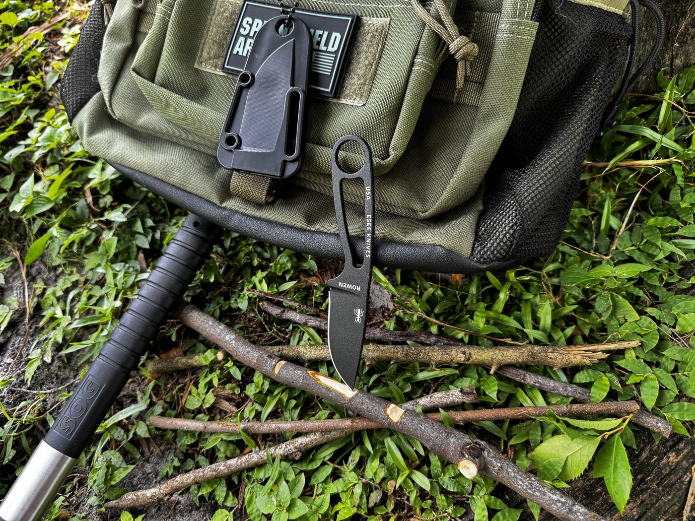 A compact fixed-blade neck knife is placed on a campsite surface next to partially whittled sticks and other outdoor gear. The knife is secured in a molded polymer sheath with a breakaway ball chain, suggesting its use as a pendant-carry tool. Nearby items such as a pack and rope emphasize a camping context. The presence of carved wood demonstrates the knife’s role in bushcraft and small camp chores like kindling prep or tool shaping. The knife’s small size makes it a lightweight addition to minimalist camping gear. This ALT text is written for visually impaired users and optimized with terms such as neck knife, camping gear, whittling, bushcraft, and fixed blade.