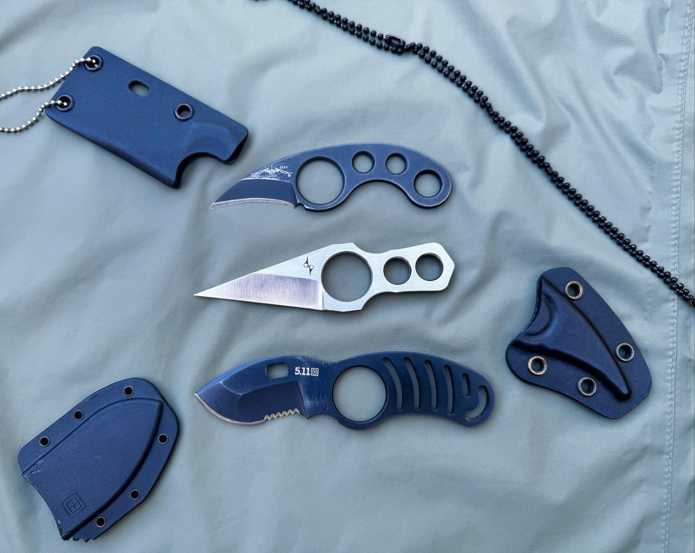 A group of compact neck knives with forward finger rings built into the handles is arranged neatly with their sheaths. The knives vary in blade shape, including drop point and claw-like profiles, but all share the circular ring that allows the user to hook a finger through for retention. This design was popularized by French knife maker Fred Perrin and remains a defining feature in many defensive and utility neck knives. Sheaths are molded polymer, suspended by short chains or cords for neck carry. The finger ring improves indexing under clothing and ensures the knife stays secure in the hand during hard use. The image introduces readers to this specialized neck knife style and its practical benefits.