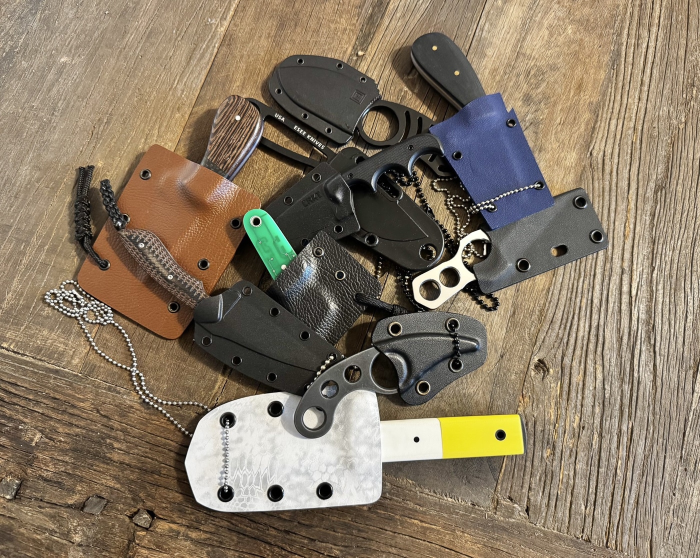 An assortment of neck knives arranged together, highlighting compact design and carry options. The blades are housed in molded polymer or Kydex sheaths with ball chain lanyards attached. Knife handles differ in size and construction, including textured scales, smooth synthetic grips, and ringed styles influenced by Fred Perrin’s La Griffe design. Blade profiles range from utilitarian drop points to aggressive Wharncliffe and hawksbill shapes. The photo emphasizes the practicality of small fixed blades carried at the sternum. It also reflects the popularity of neck knives for water sports, bushcraft, and self-defense. This general view introduces readers to the range of neck knife models available.