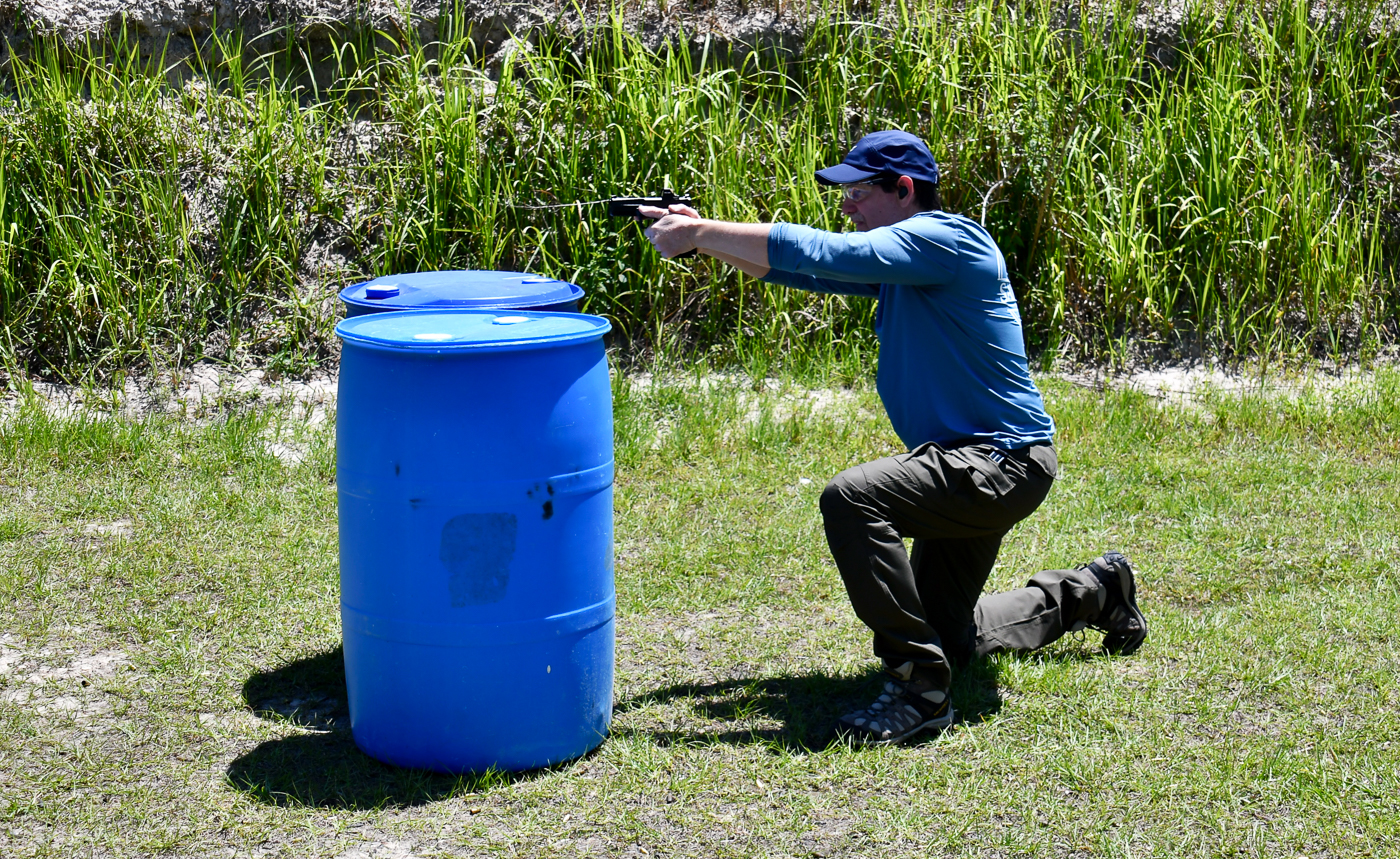 A shooter is shown with his pistol slide locked back during a live-fire training session, prompting an immediate reload behind cover. He leans into a controlled reload while keeping his muzzle pointed downrange and eyes on the threat area. The photo captures the critical moment when a stoppage interrupts the flow of action and the shooter must perform a reload or clearance. This scene ties into two self-defense shooting drills that emphasize movement, use of cover, and managing malfunctions. The environment includes paper targets and simulated cover, showing a realistic training bay. The image underscores the importance of practicing reloads and saved-mag techniques with an EDC handgun to stay ready under stress.