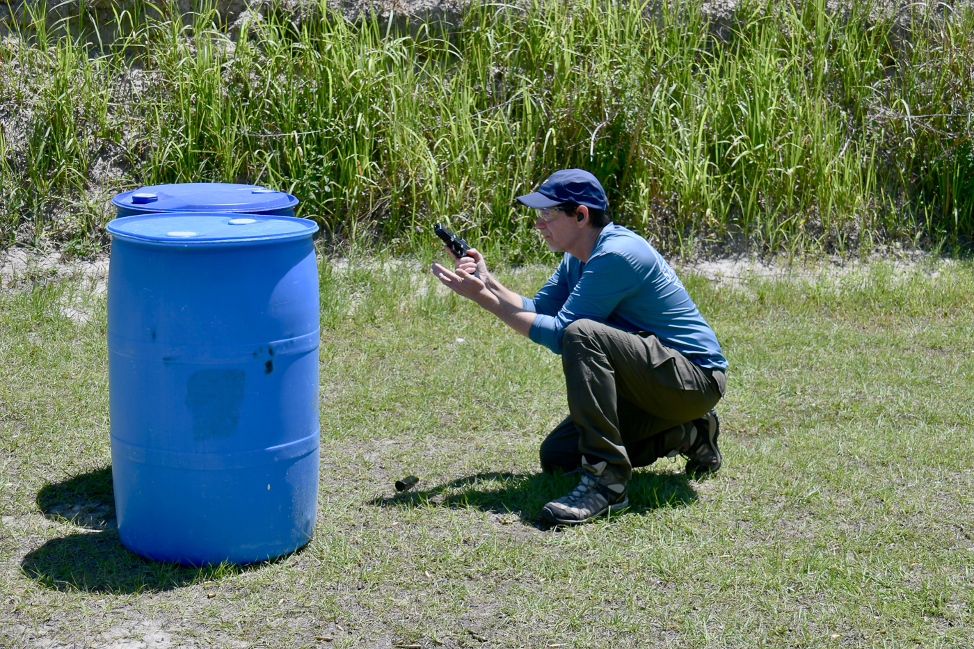 A man is shown at an outdoor shooting range using a large plastic drum as cover while reloading his Springfield Armory pistol. He keeps his body low and the muzzle pointed downrange, minimizing exposure to the simulated threat. The image captures a moment in tactical practice where movement, cover use, and reload technique come together. This scene is part of two self-defense shooting drills that emphasize minimizing risk while getting the firearm back into the fight. Background elements include paper targets and an open training bay that suggest a controlled environment. The photograph illustrates responsible range behavior, focusing on muzzle discipline, situational awareness, and the practical use of improvised cover.