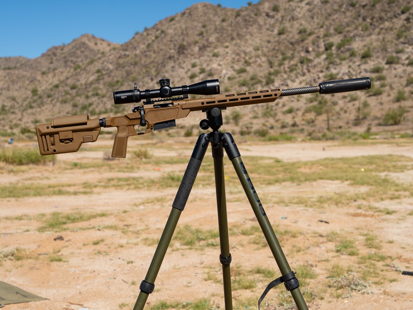 Precision rifle shooter engaging targets from supported tripod position with Springfield Heatseeker chassis rifle in 6.5 Creedmoor caliber. The carbon fiber barreled rifle clamped into BOG Deathgrip tripod head provides rigid platform for accurate shot placement at distance. Shooter demonstrates proper form with eye aligned to riflescope and support hand controlling rear bag or stock adjustment. Suppressor visible on muzzle helps manage recoil and protects hearing during extended shooting sessions. This setup used successfully for first-round impacts on 10-inch steel plates out to 500 yards with both match and hunting ammunition. Tripod shooting position allows for more flexible shooting angles compared to prone bipod work. The lightweight chassis design of Model 2020 makes it well-suited for this type of positional shooting.