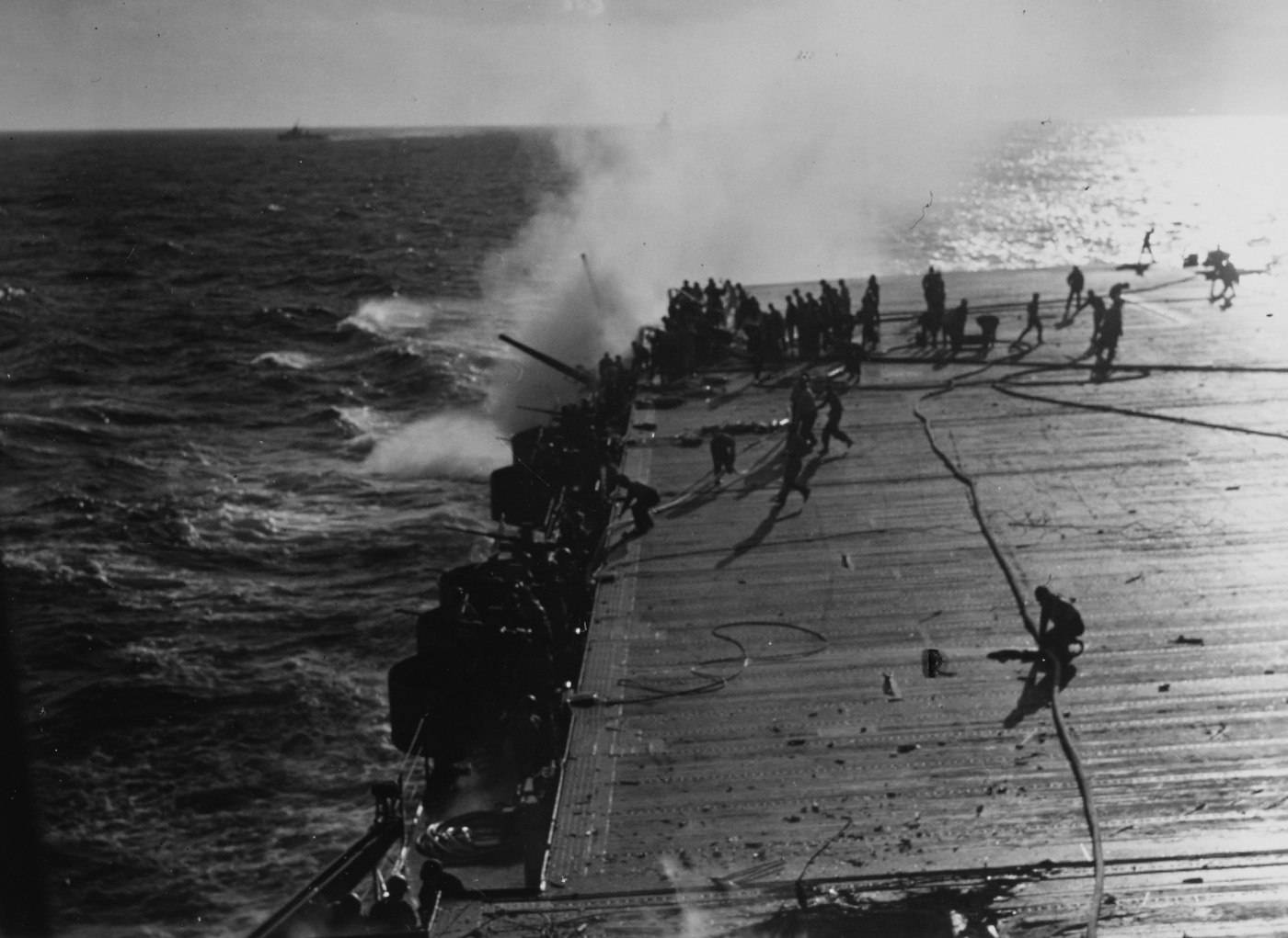 This photo shows sailors aboard USS Enterprise (CV-6) fighting a fire on the flight deck after the carrier was hit by a Japanese bomb during the Solomon Islands campaign in 1942. Thick smoke billows upward as crew members operate hoses and carry firefighting equipment across the deck. Flames are visible in the damaged section, with debris scattered from the impact. Other sailors are seen assisting wounded or moving ammunition to safety. The intensity of the scene highlights the chaos and urgency of damage control in battle. Despite severe damage, the crew’s rapid response kept the fire from spreading and allowed Enterprise to remain operational. The image documents both the vulnerability of even major warships and the effectiveness of well-trained naval teams under fire. It’s a striking moment from one of the carrier’s toughest engagements.