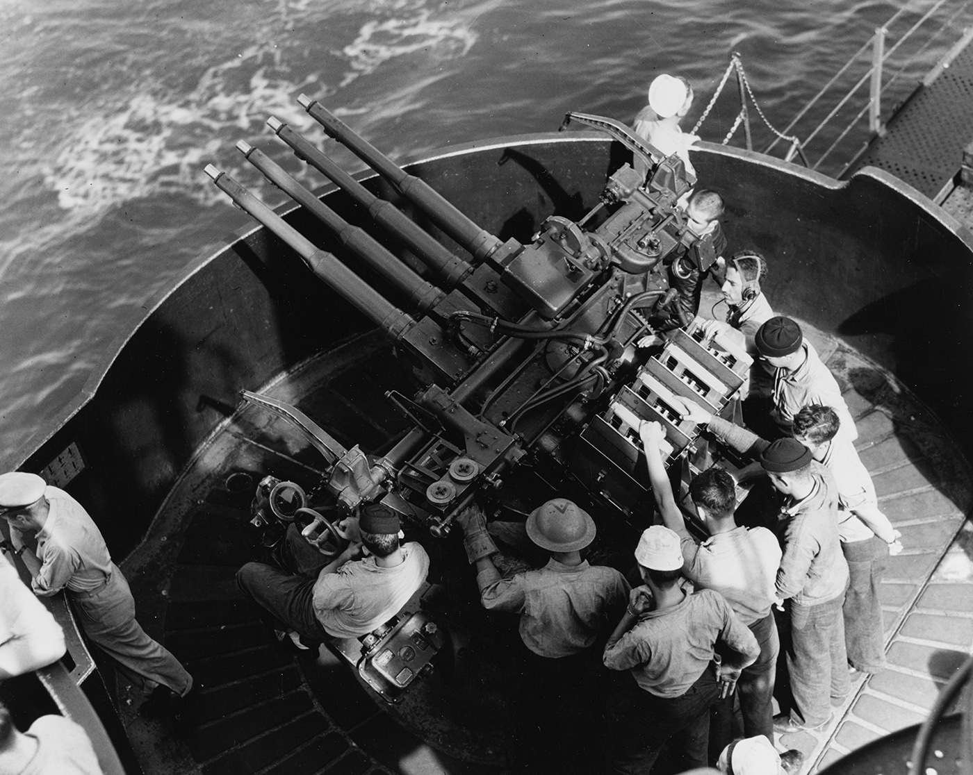 This photo shows sailors on USS Enterprise (CV-6) manning anti-aircraft (AA) guns during the Wake Island raid in World War II. The gun crews are positioned at their weapons, wearing helmets and protective gear as they scan the sky for enemy aircraft. Each sailor is focused on his assigned position, with hands on controls and ammunition nearby. The background reveals parts of the flight deck and the open ocean, highlighting the exposed nature of AA gun stations. The guns are aimed skyward, ready to respond to any Japanese air attack that might threaten the ship or its aircraft. The tension and readiness of the crew are apparent, reflecting the real danger faced during every carrier raid. Defensive positions like this were a critical line of protection for the ship throughout the Pacific campaign.
